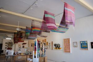 A gallery space with white walls with paintings hanging, as well as brightly colored fabric draped, hanging from the ceiling 