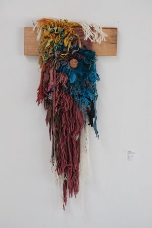 A fiber art sculpture made of yarn and scraps of fabric in a rainbow of colors on a white wall