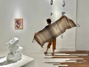 A woman wearing ochre colored overalls wears a brown, tan, white gradation shawl in a white and wooden gallery space