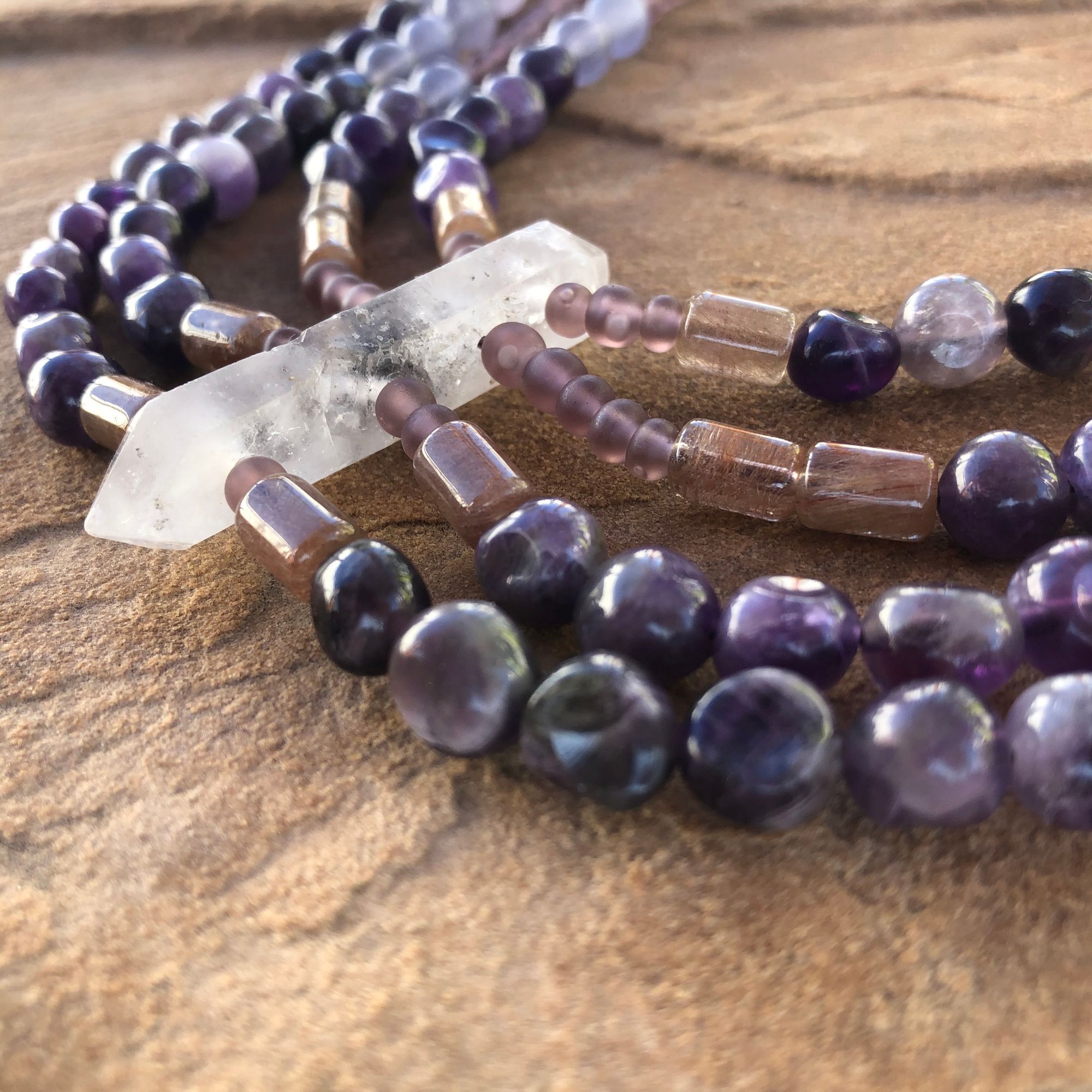 purple amethyst necklace with pink rutilated quartz, purple glass and clear quartz beads laying flat on a tan stone