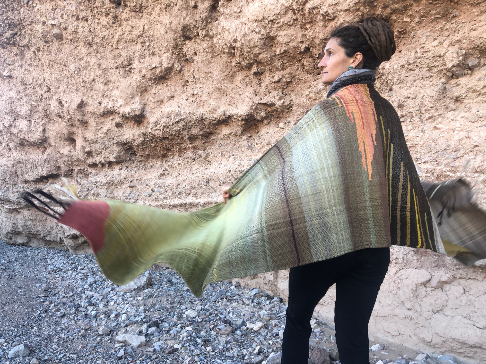 woman standing in the desert with her arms outstretched wearing a handwoven naturally dyed brown, green, yellow, grey, red and salmon shawl.