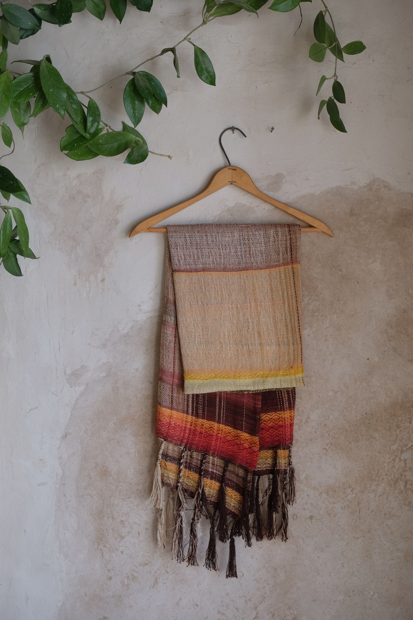 Handwoven red, orange, yellow, brown and white scarf on a hanger on a white mud wall