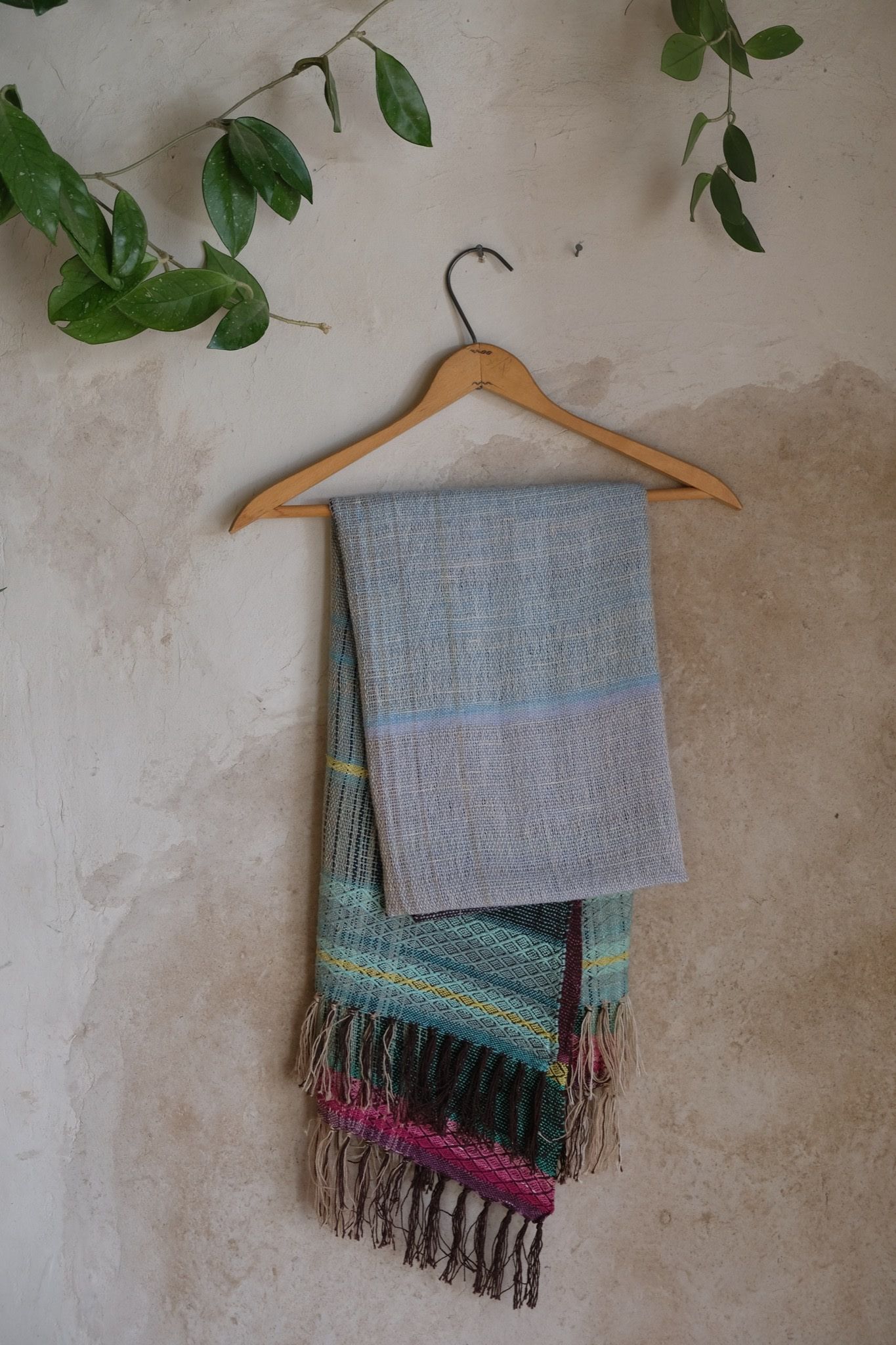 Blue, pink, yellow, purple and teal handwoven silk scarf on a hanger hanging on a white mud wall