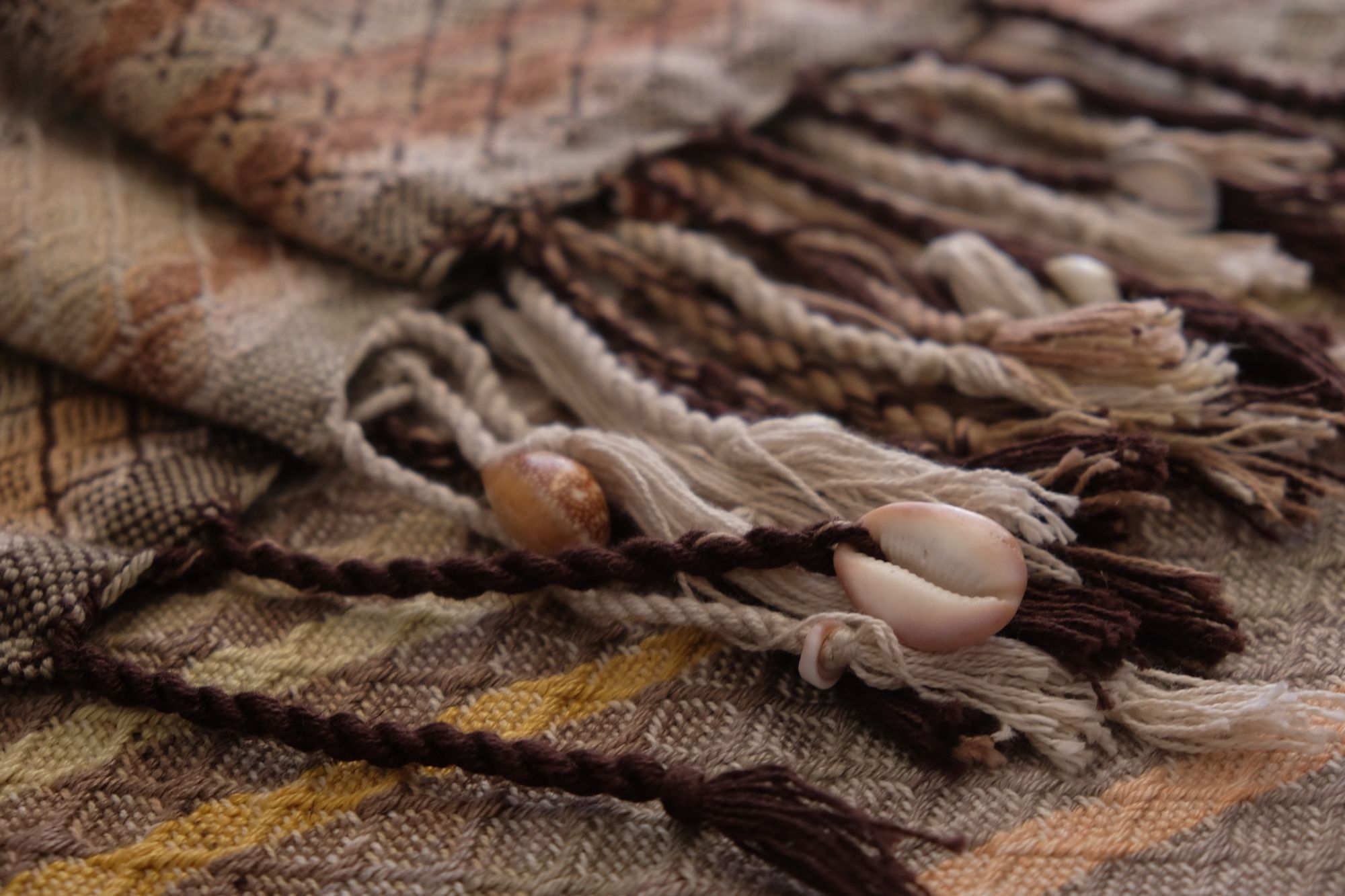 Detail of a handwoven pink, brown, tan, yellow and grey shawl with diamond pattern weave with long twined fringe and shells tied into it