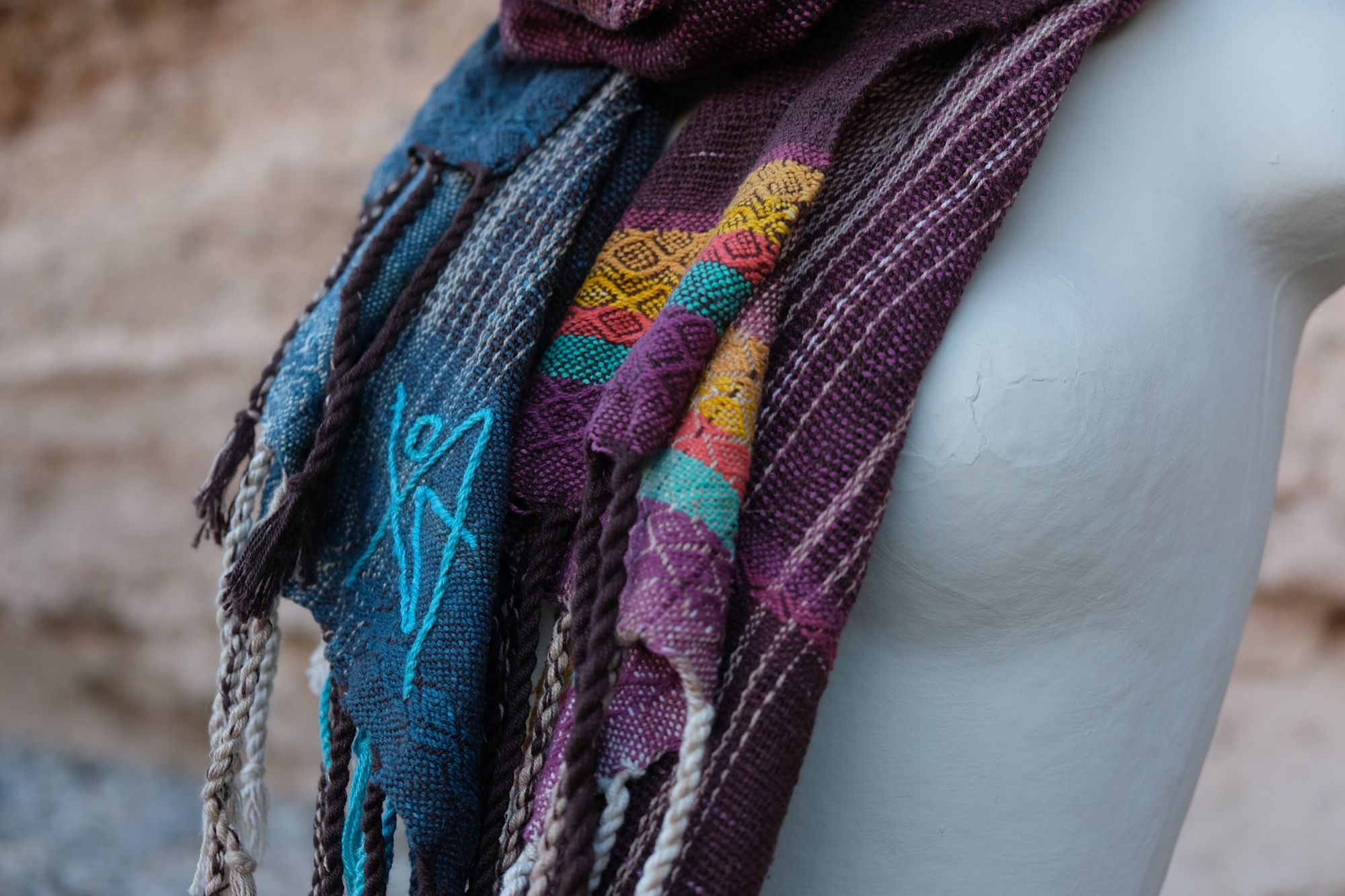 Handwoven maroon, blue, yellow and teal scarf on a white mannequin sitting on a pile of rocks