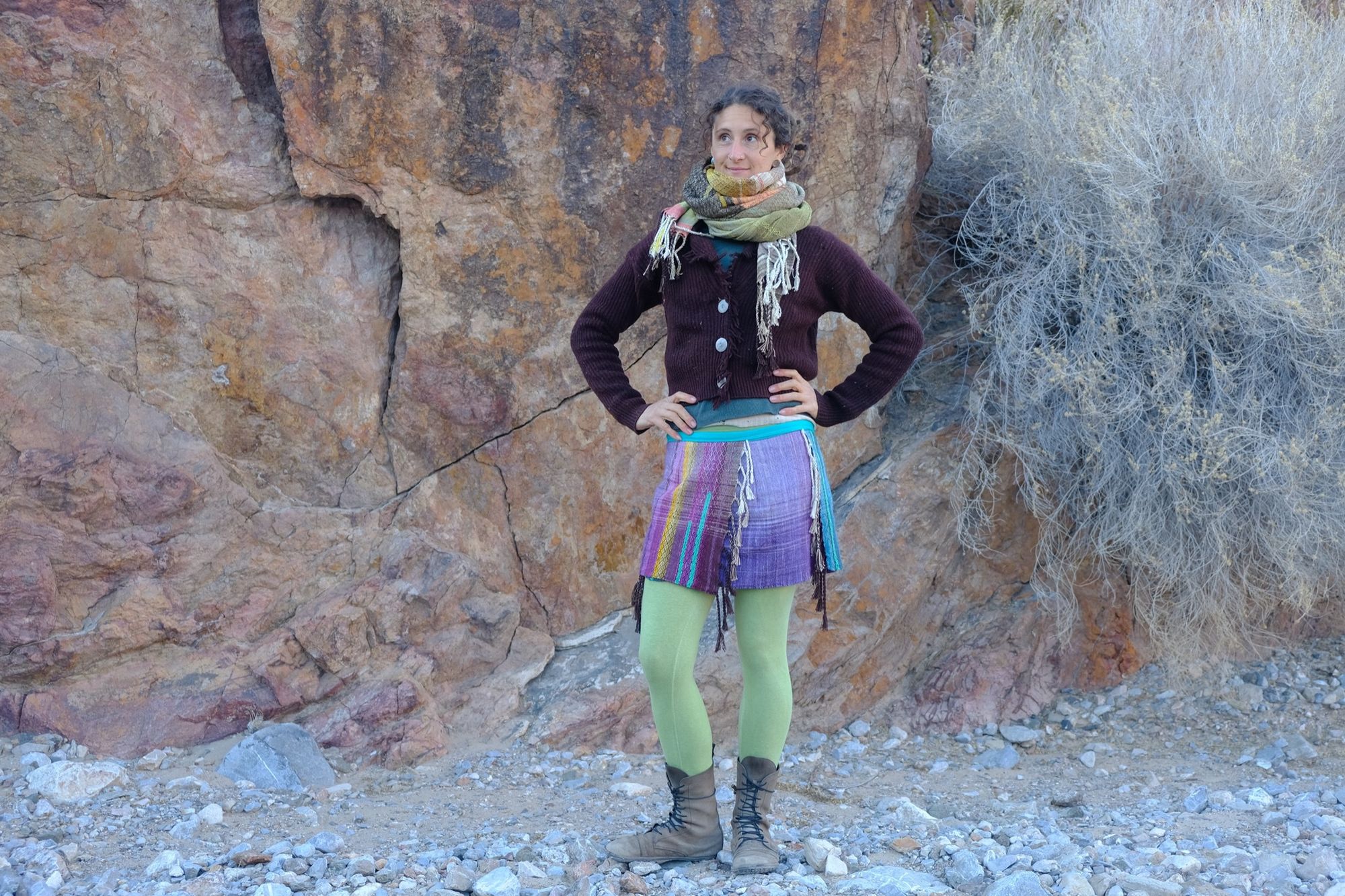 Woman wearing blue and purple fringed skirt with green leggings, a brown sweater and large scarf against a stone wall