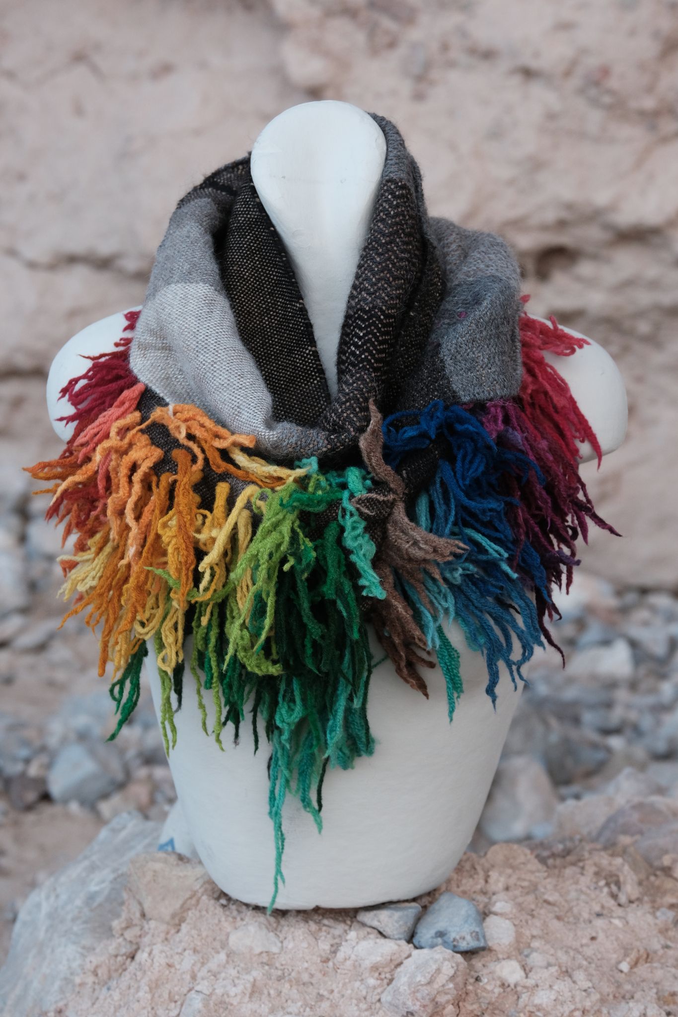 Highly textured scarf in greyscale with rainbow fringe like a lions mane on a white mannequin with a tan background 