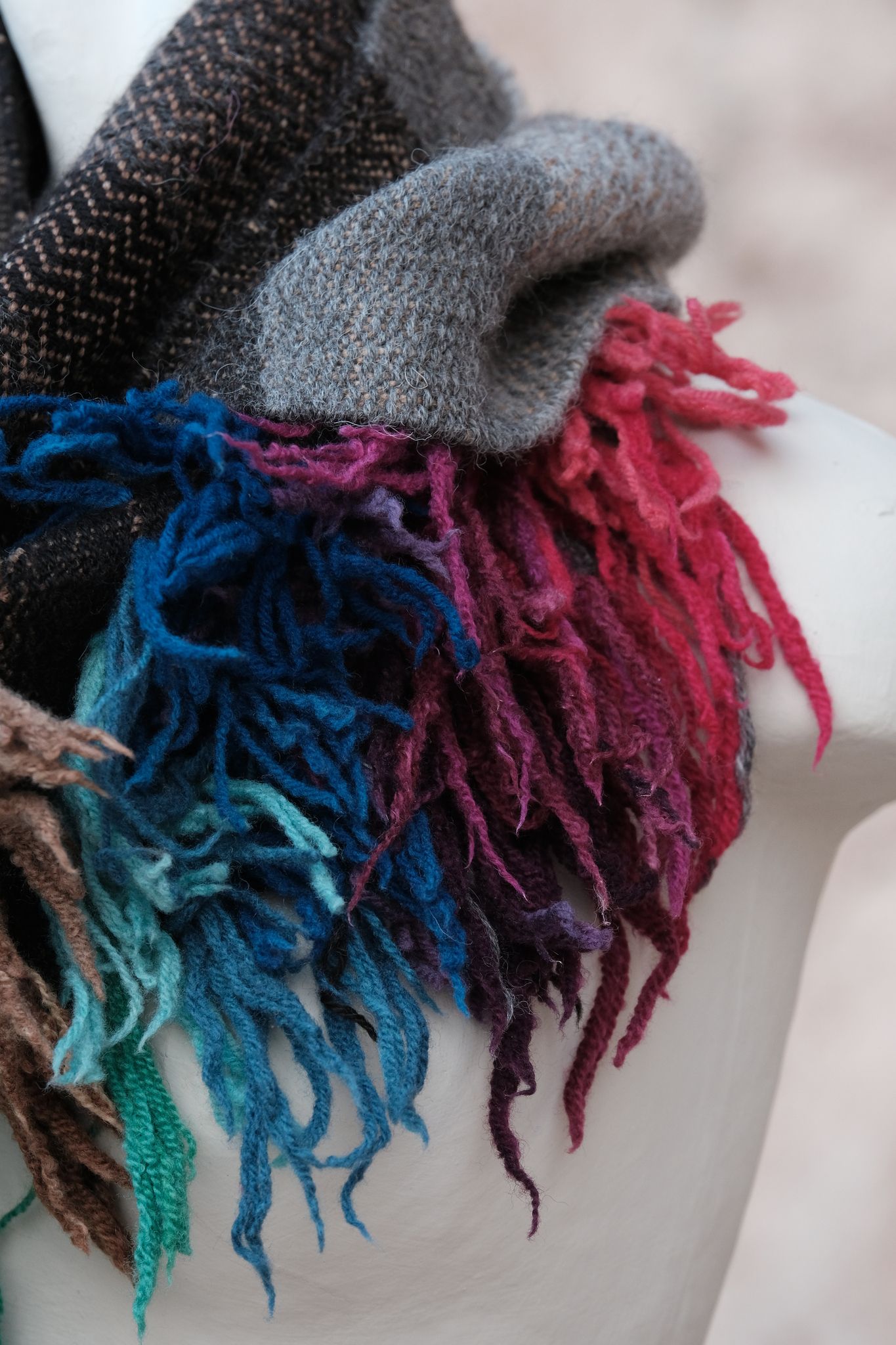 Highly textured scarf in greyscale with rainbow fringe like a lions mane on a white mannequin with a tan background 