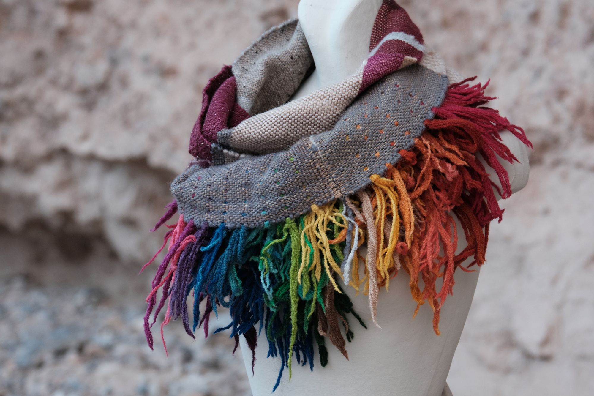 Rainbow and grey sculptural fringed scarf on a white mannequin sitting on tan stones with an earthen tan background