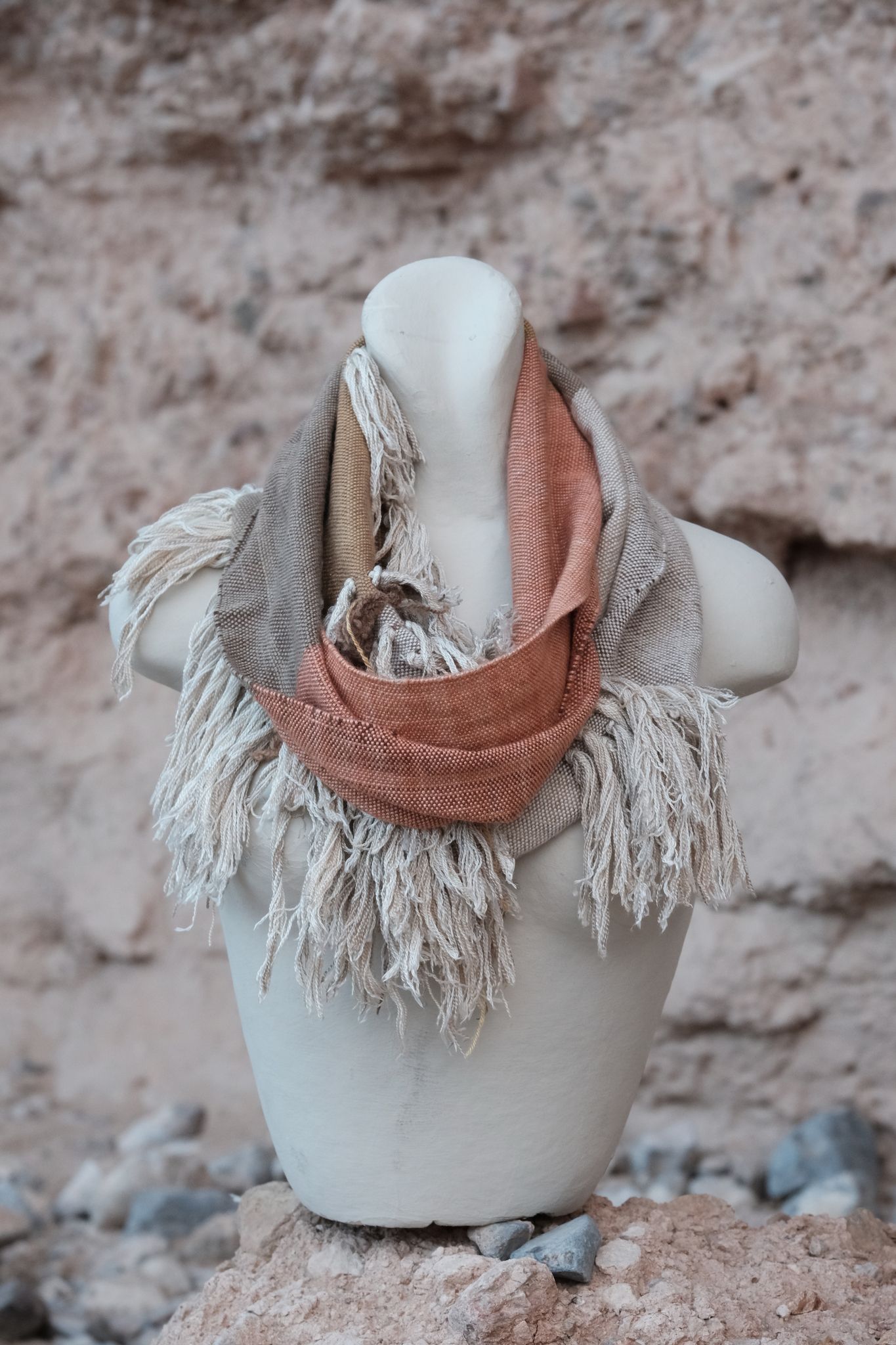 salmon colored, brown, white and grey scarf with white cashmere fringe on a white mannequin sitting on a tan rock. 