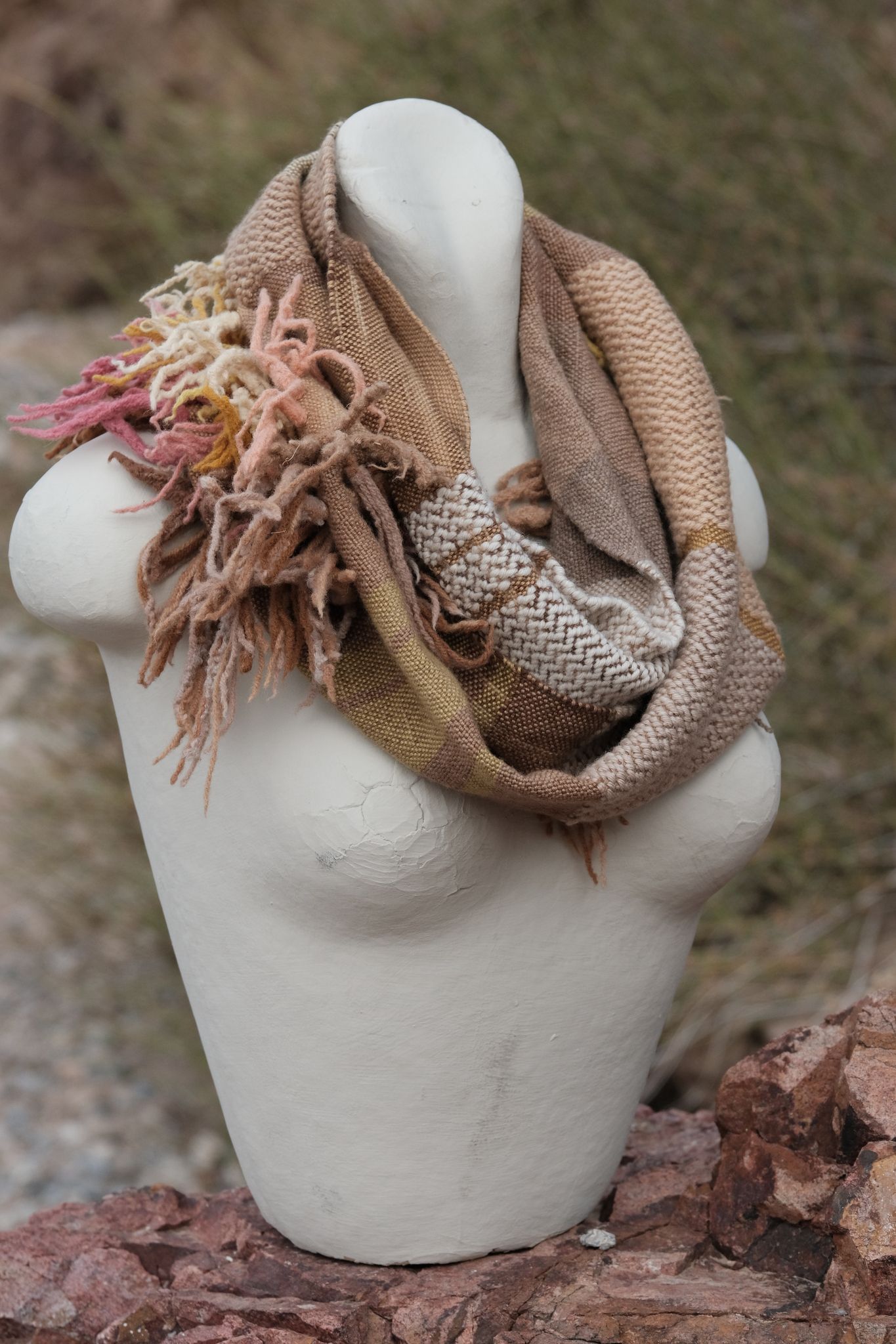 Handwoven naturally dyed brown, pink, yellow, tan and red merino scarf with sculptural fringe on a white mannequin, sitting on a tan rock.