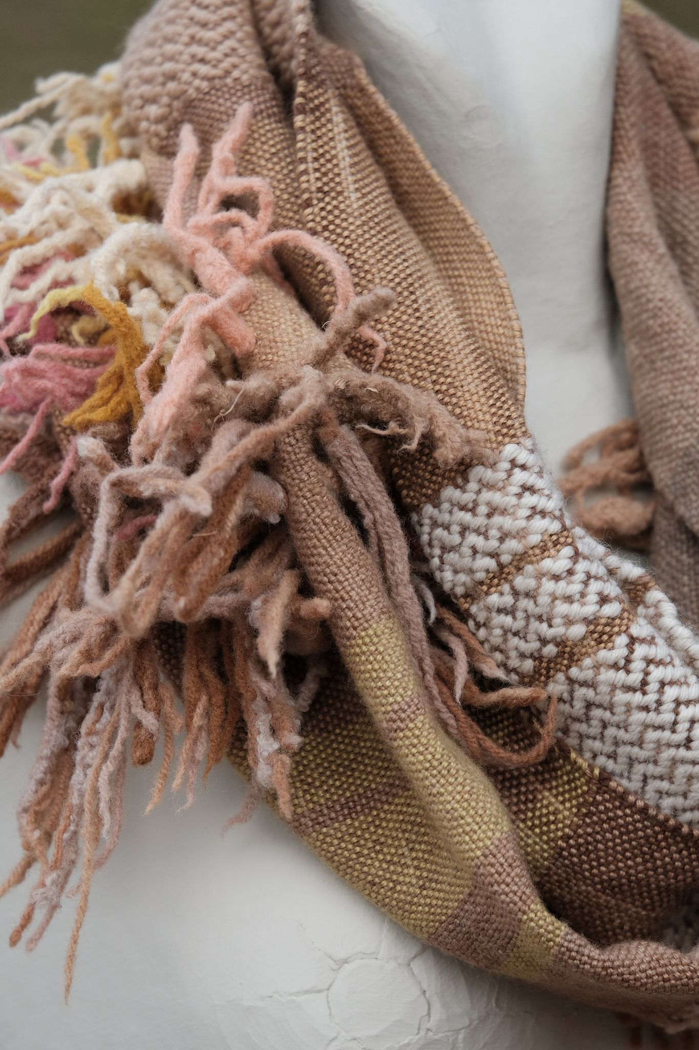 detail of Handwoven naturally dyed brown, pink, yellow, tan and red merino scarf with sculptural fringe on a white mannequin, sitting on a tan rock.