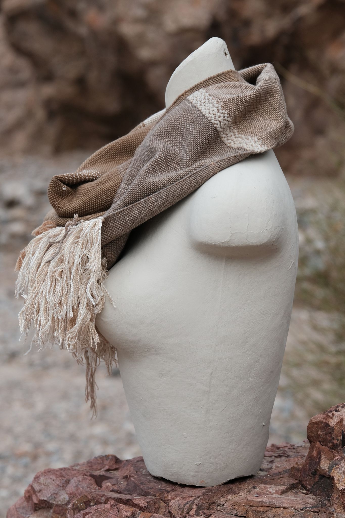 Handwoven naturally dyed brown, grey, yellow, pink and green scarf with sumptuous sculptural fringe on a white mannequin on a rock in the desert
