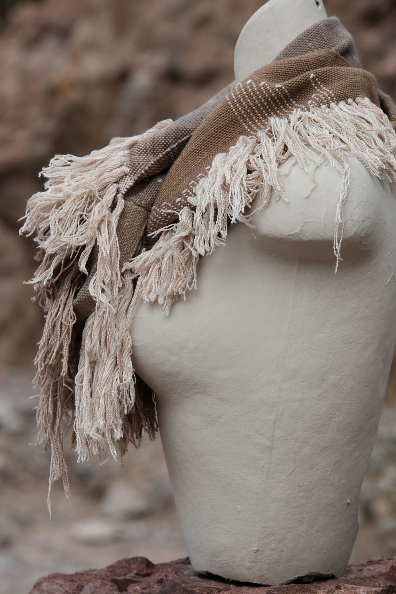 Handwoven naturally dyed brown, grey, yellow, pink and green scarf with sumptuous sculptural fringe on a white mannequin on a rock in the desert