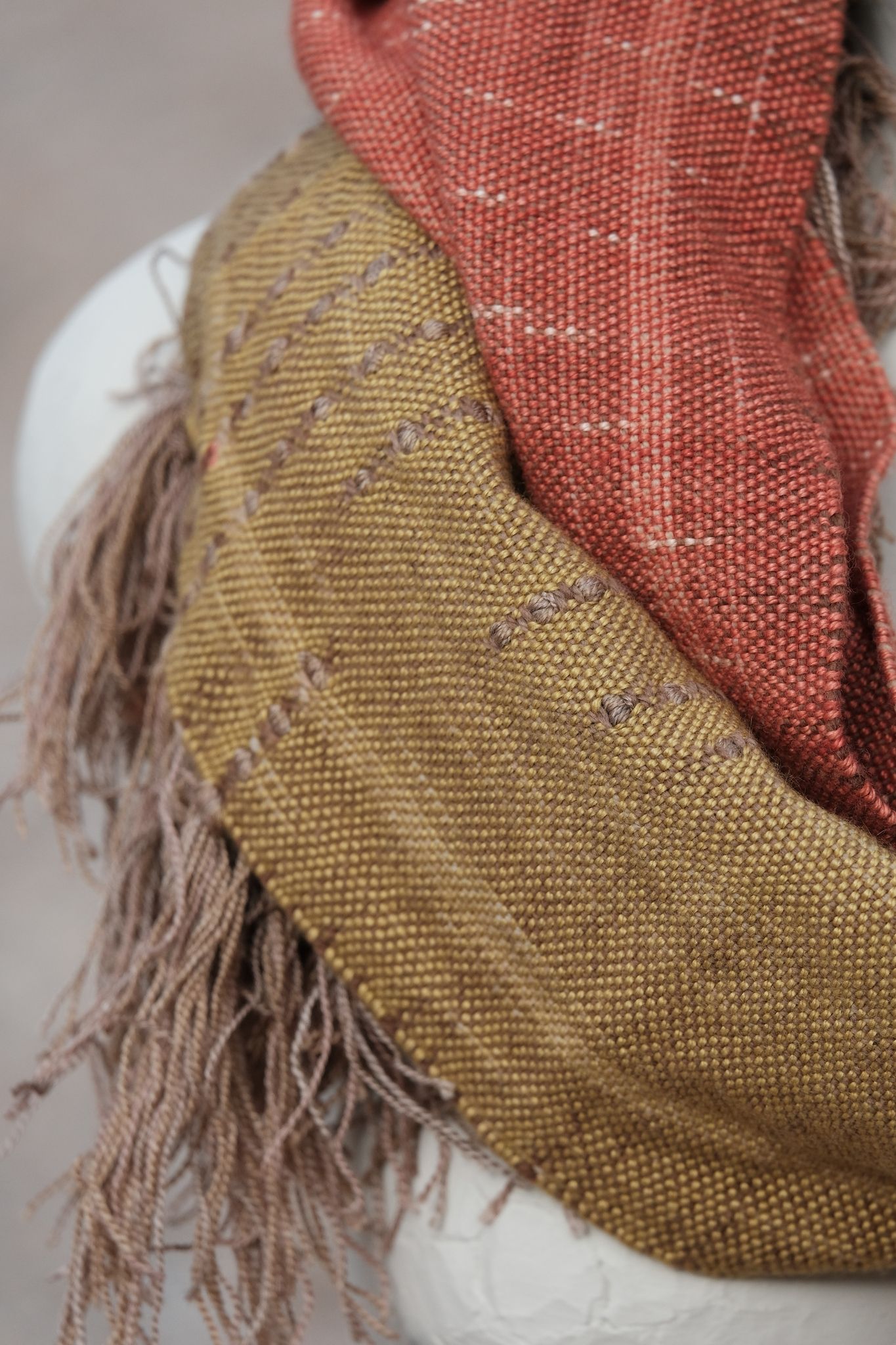 handwoven highly textured scarf that is mustard, brown, grey-blue and salmon colored, on a white mannequin sitting on a rock