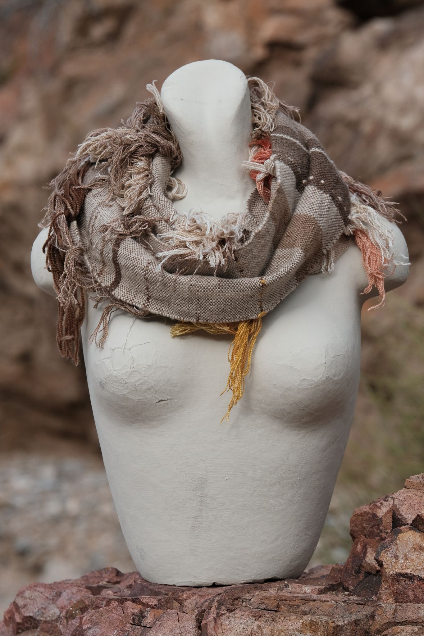 Grey and white handwoven cashmere scarf with naturally dyed salmon, brown and yellow fringe on a white mannequin sitting on a rock in the desert