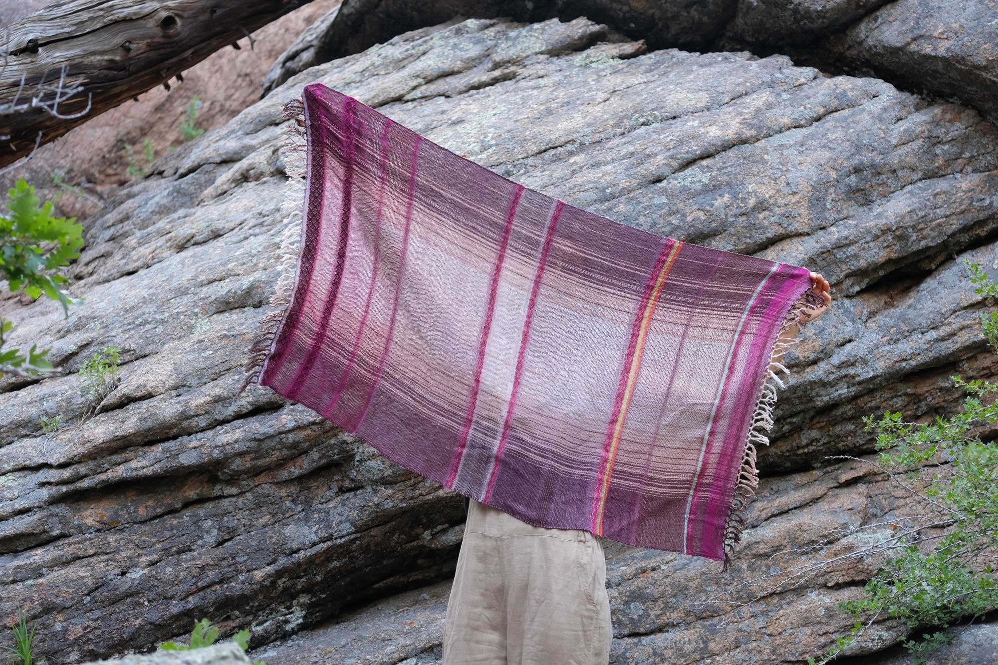 Woman wearing brown overalls and handwoven raspberry pink, brown and white scarf standing in front of a boulder