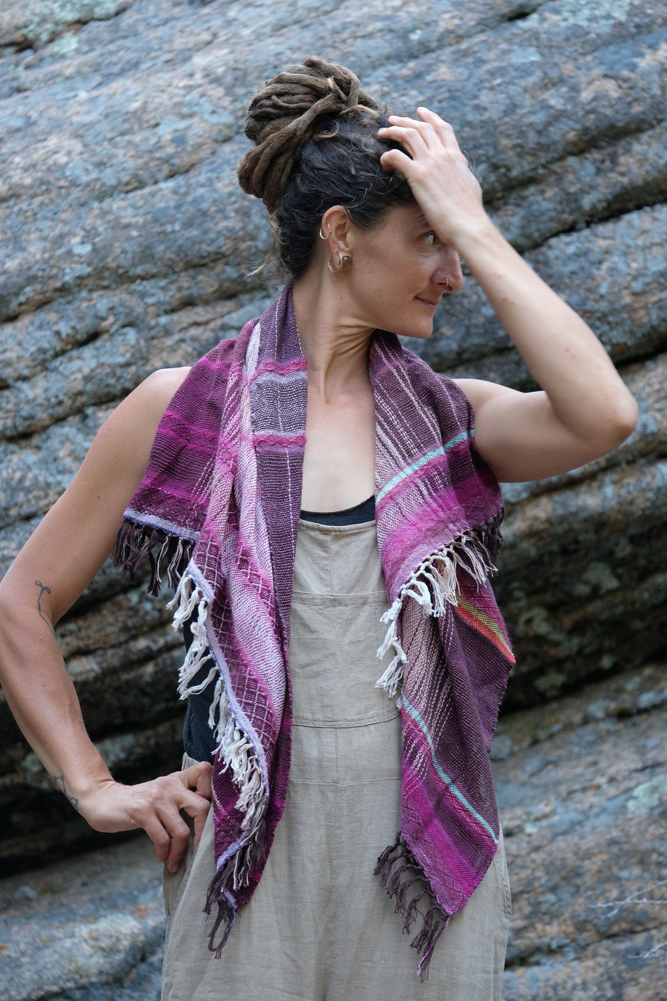 Woman wearing brown overalls and handwoven raspberry pink, brown and white scarf standing in front of a boulder