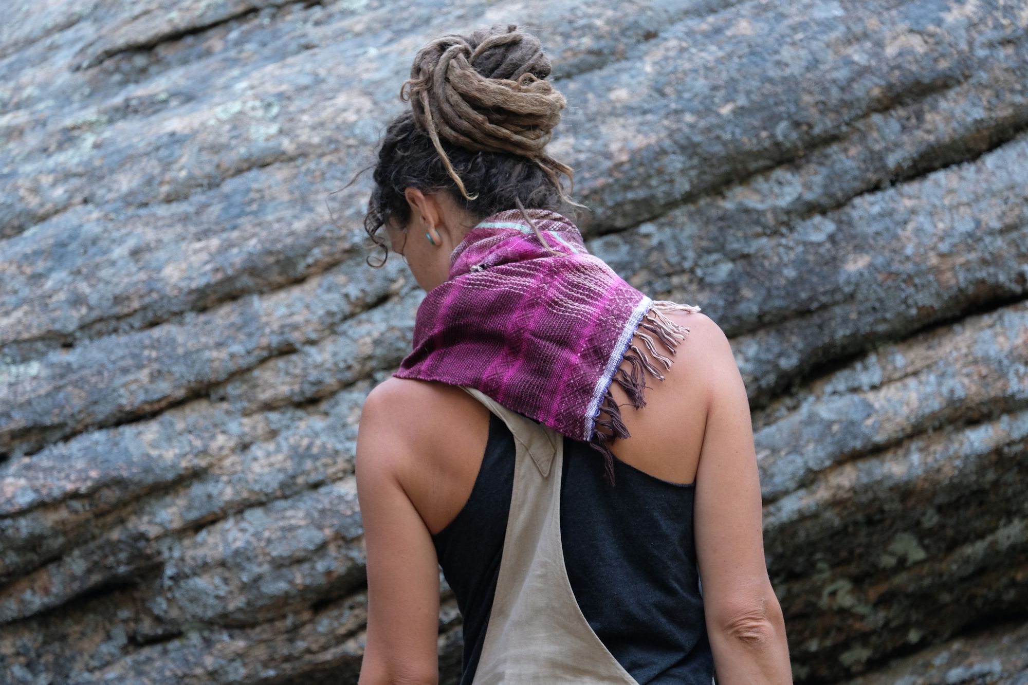Woman wearing brown overalls and handwoven raspberry pink, brown and white scarf standing in front of a boulder