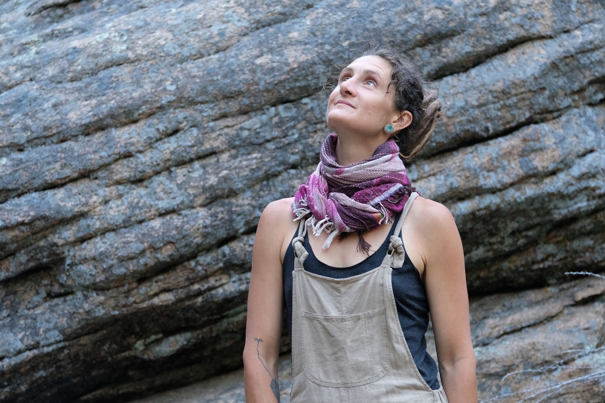 Woman wearing brown overalls and handwoven raspberry pink, brown and white scarf standing in front of a boulder