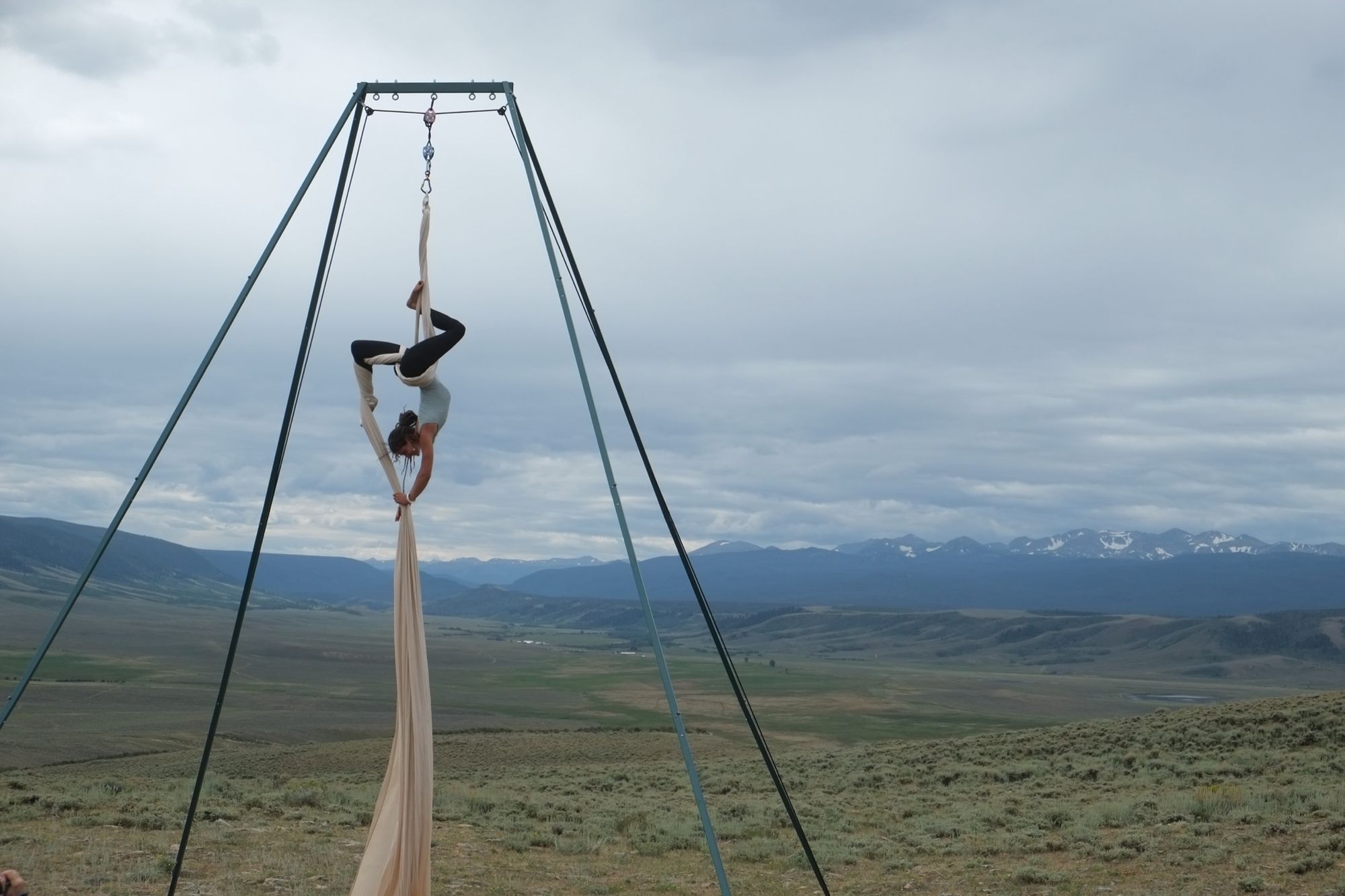 Woman wearing black pants and blue shirt doing aerial acrobatics with white fabric in front of a dramatic green mountain landscape