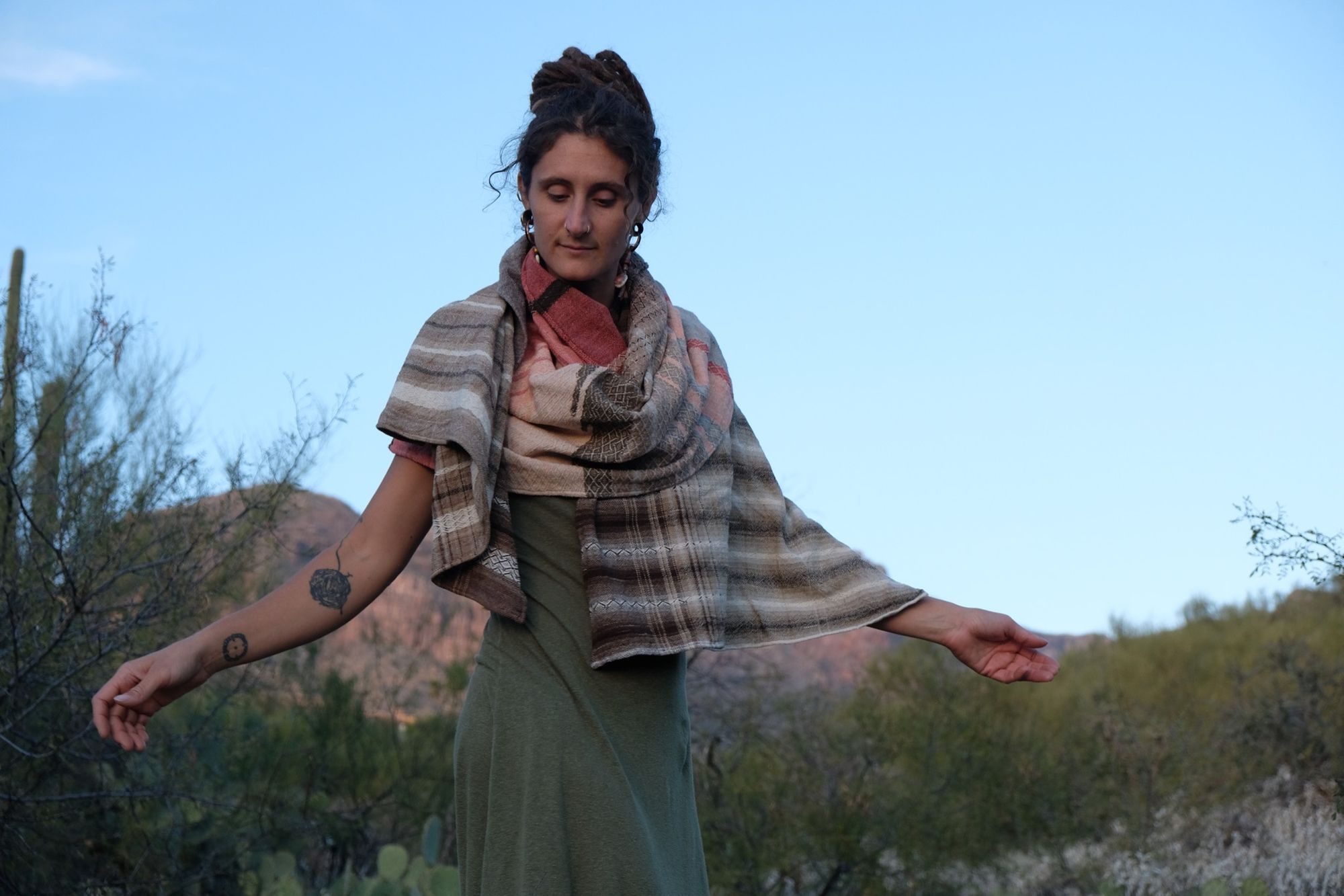 Woman in a green dress wearing a brown and salmon shawl in the desert with mountains and cactus in the background. 