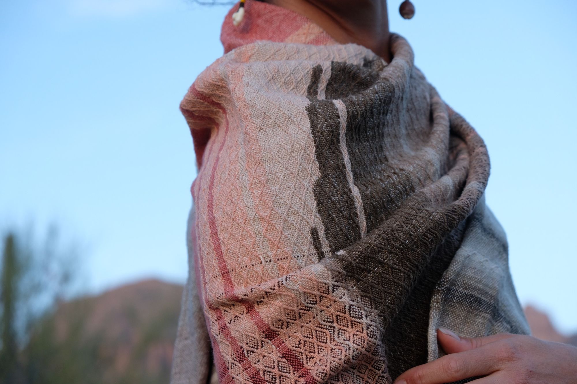 Woman in a green dress wearing a brown and salmon shawl in the desert with mountains and cactus in the background. 