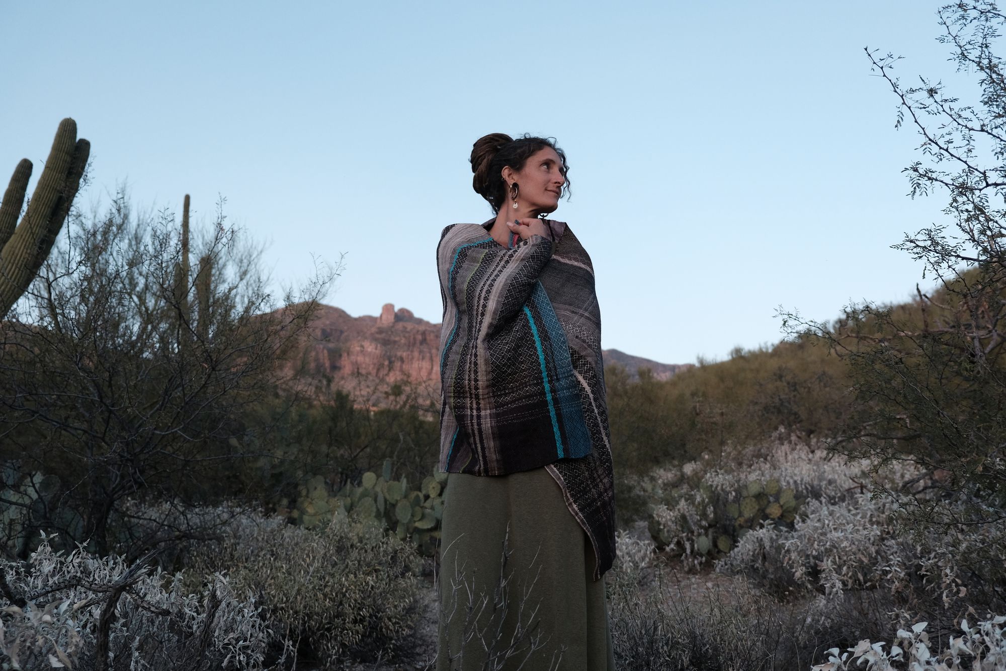 Woman wearing a handwoven black, pink, blue and lavender shawl with a crescent moon on it, standing in a desert landscape