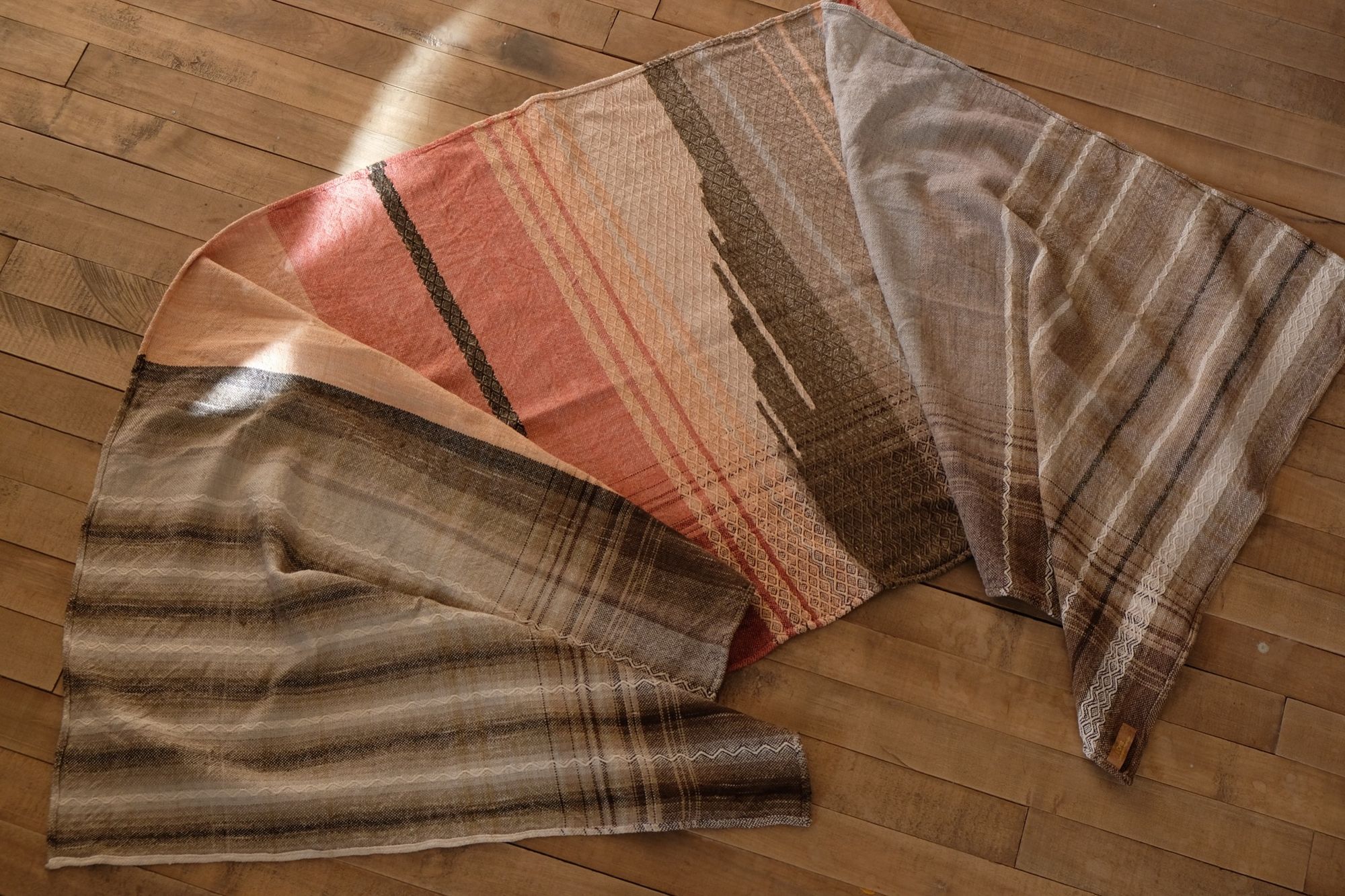 Handwoven shawl in brown, tan and salmon pink colors laying on a wood floor. 