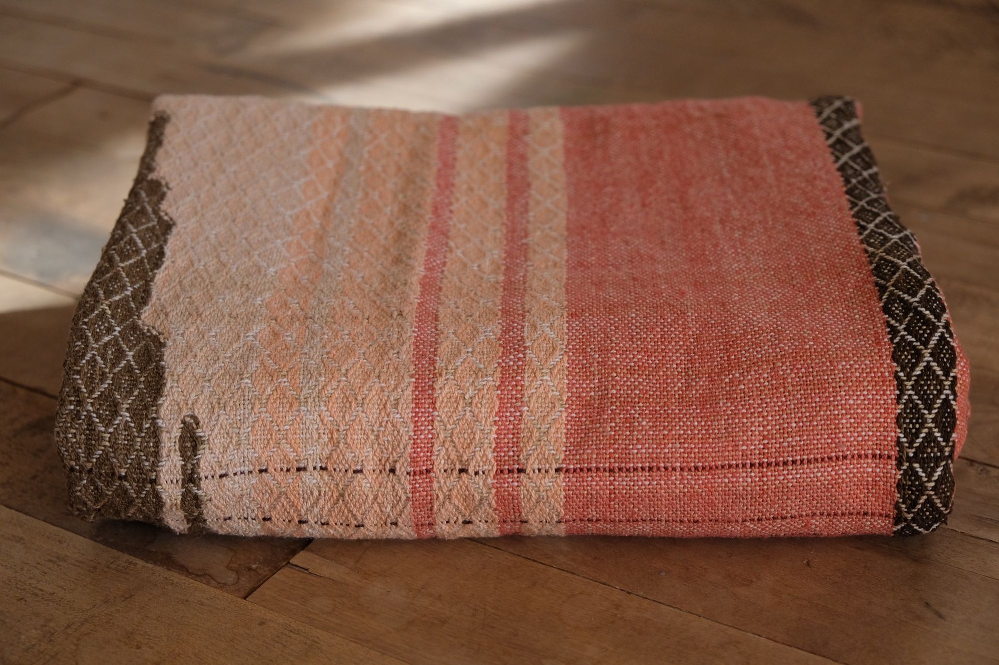 Detail of Handwoven shawl in brown, tan and salmon pink colors laying on a wood floor. 