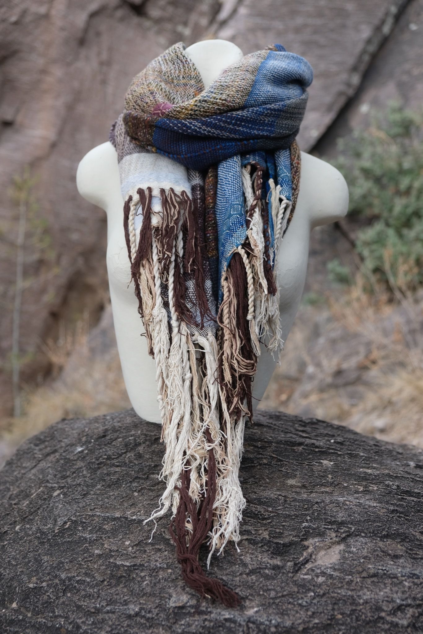 Blue, white and brown handwoven scarf on a white mannequin sitting on a boulder.