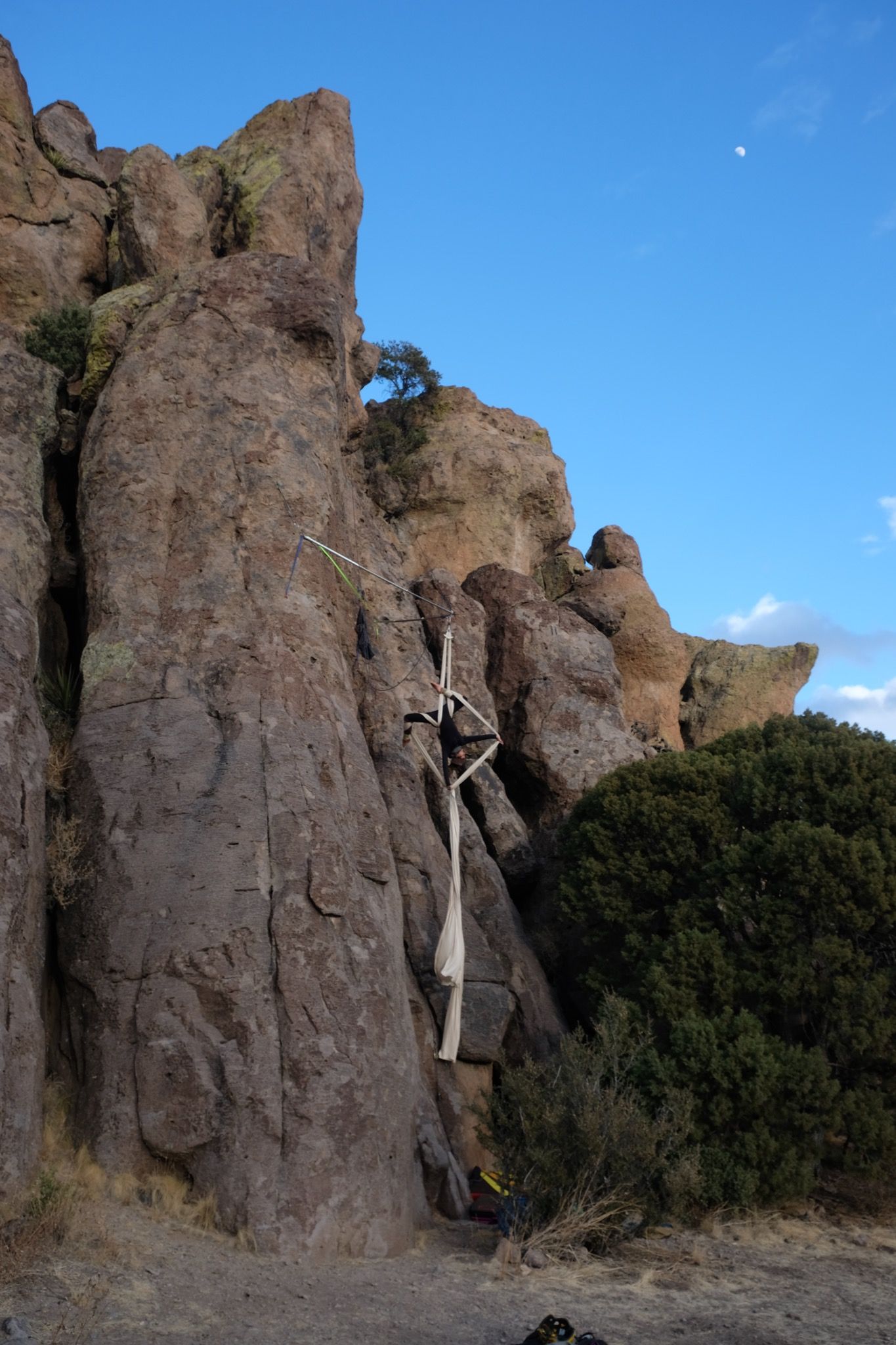 Woman hangs off apparatus, performing aerial silks with white fabric, off cliff face