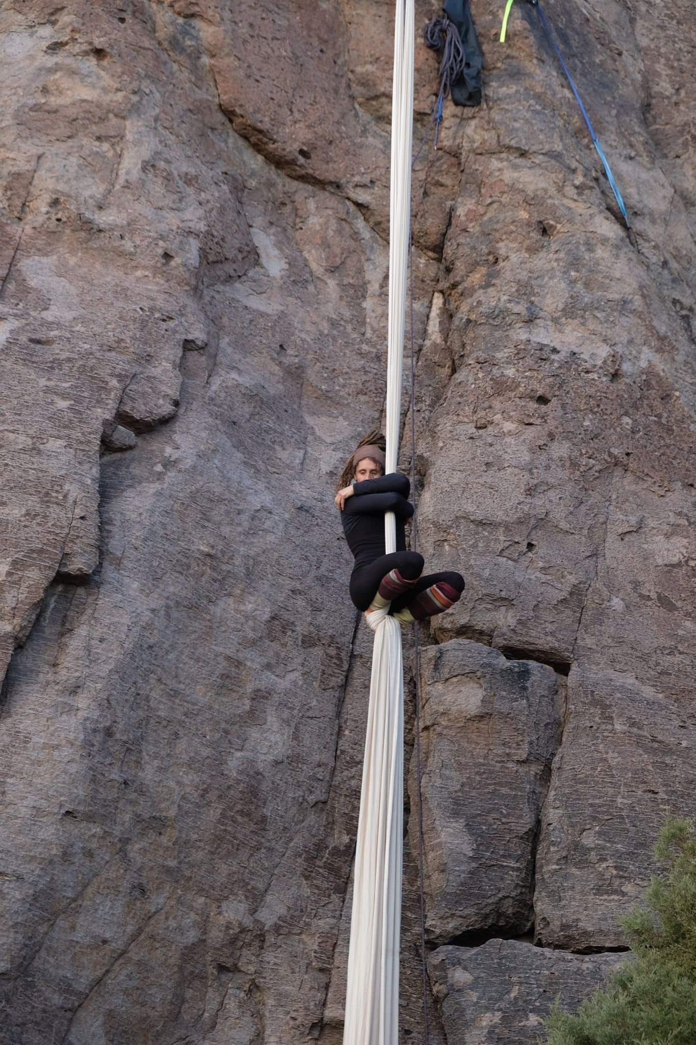 Woman hangs off apparatus, performing aerial silks with white fabric, off cliff face