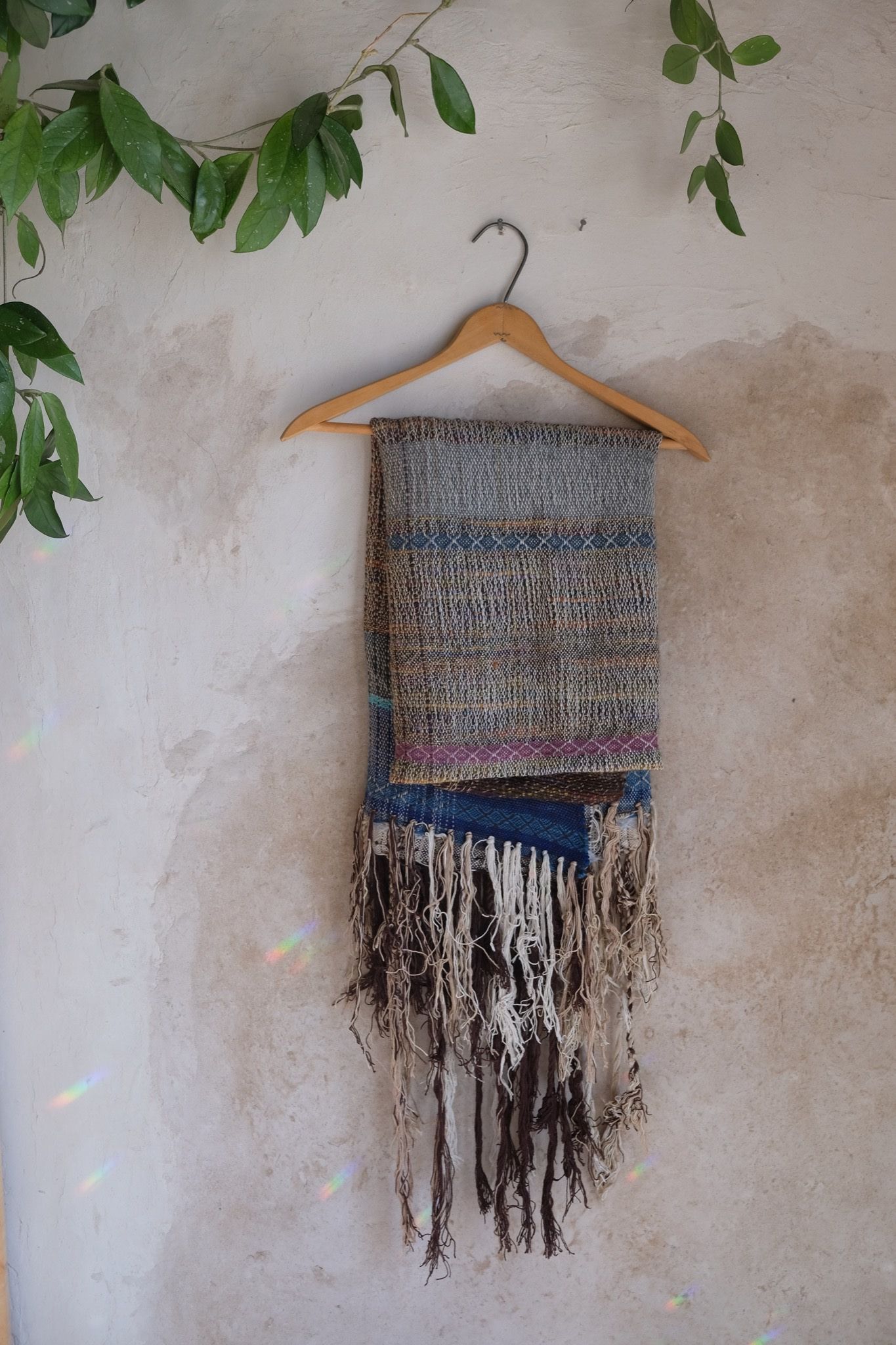 Blue, white and brown handwoven scarf on a hanger hanging on a white mud wall.