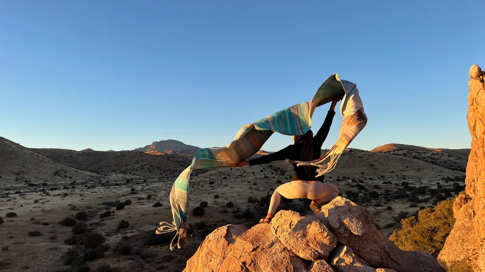 Woman standing on a cliff-top in desert mountain landscape at sunset, she is holding blue fabric which is blowing in the wind