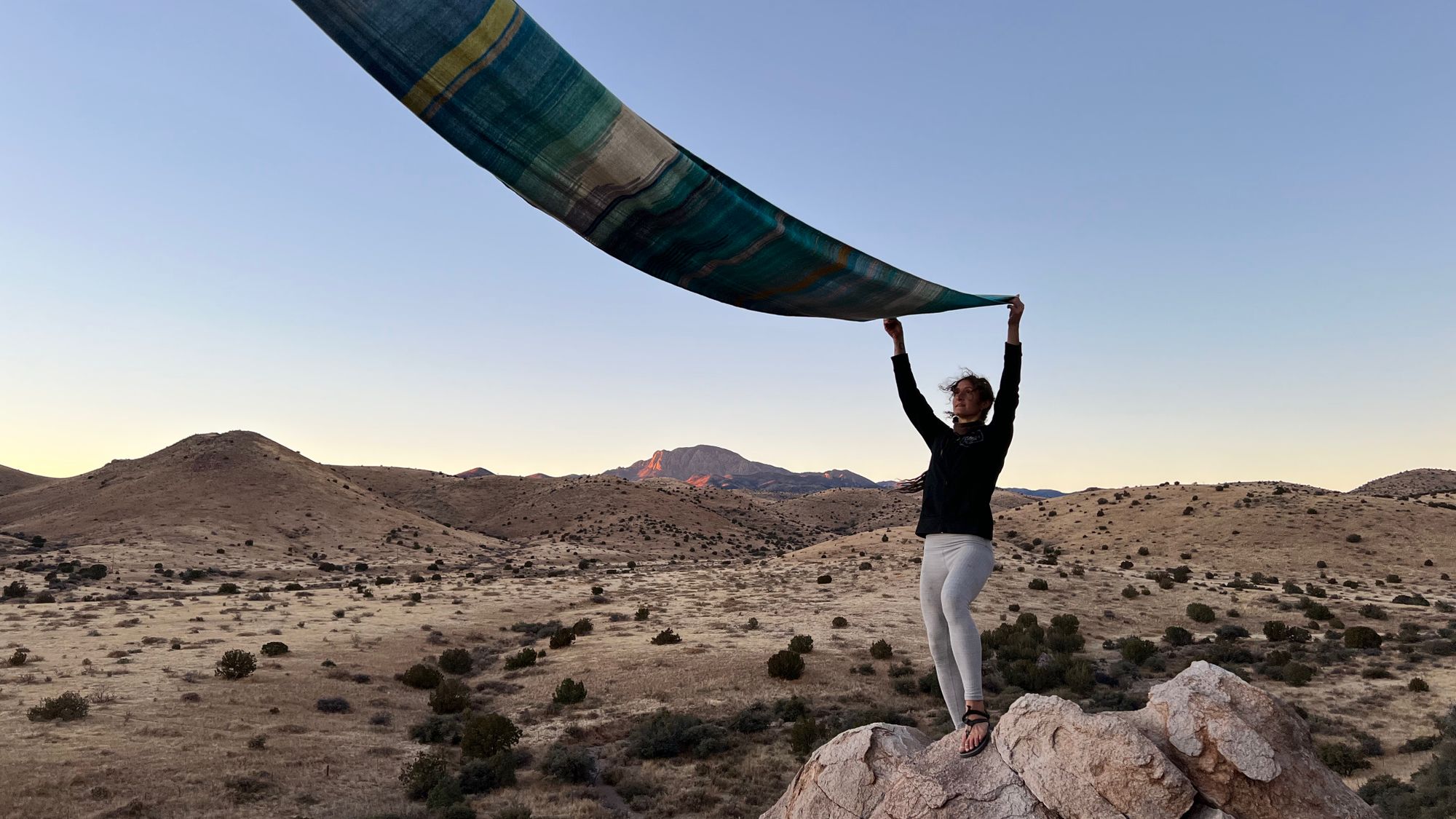 Woman standing on a cliff-top in desert mountain landscape at sunset, she is holding blue fabric which is blowing in the wind