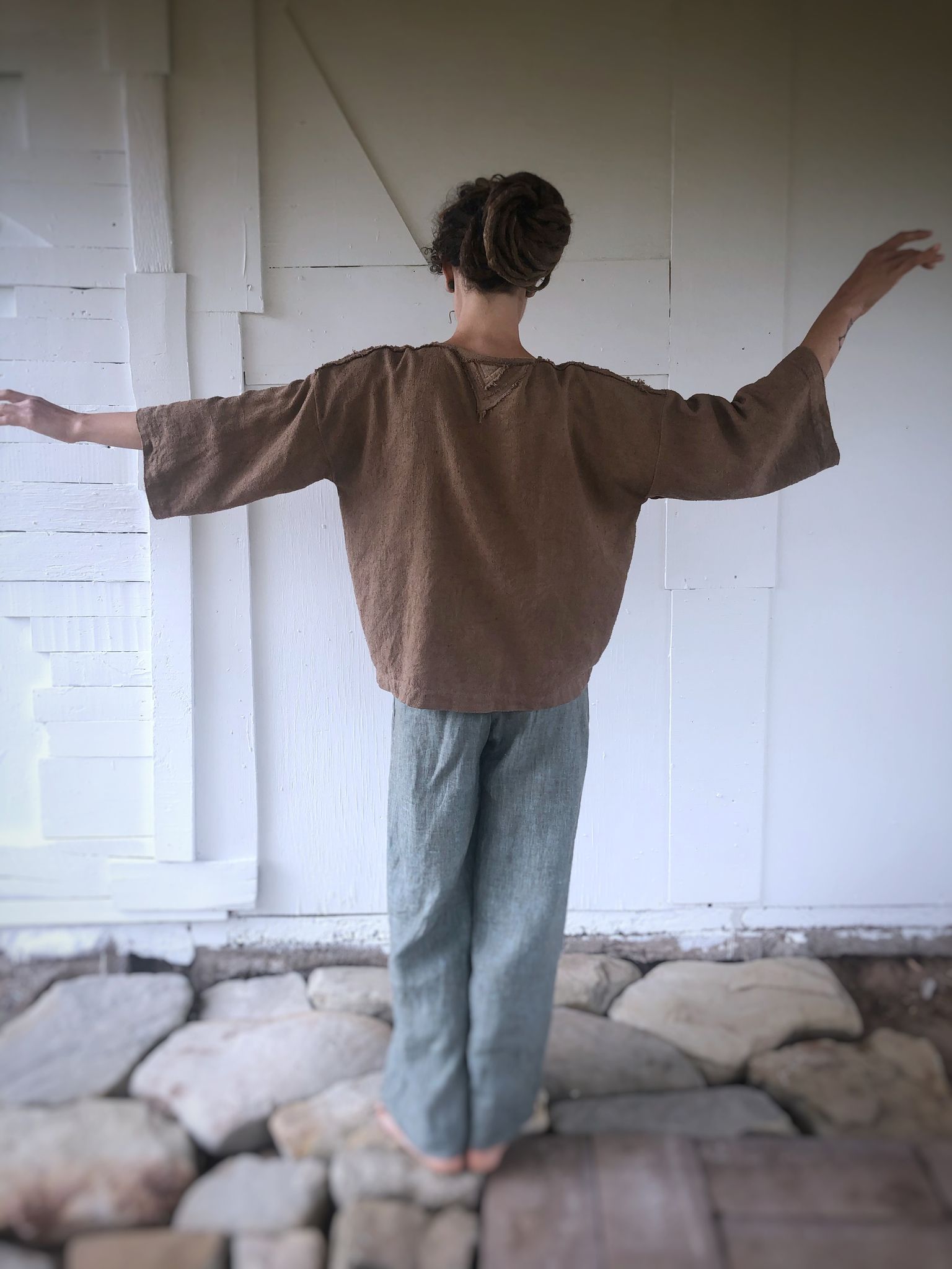 woman wearing handmade brown jacket and blue pants, standing in front of a white wall