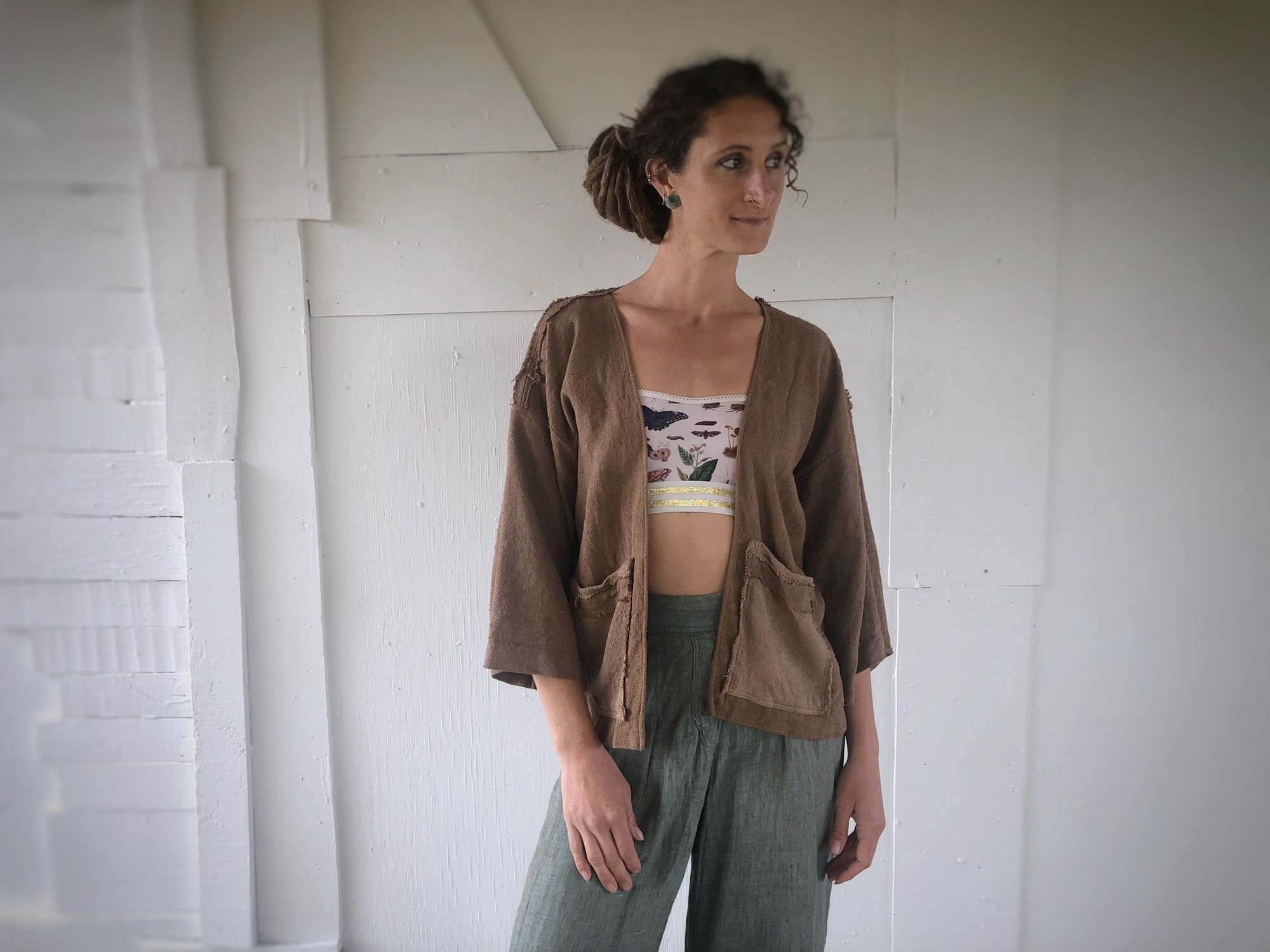 woman wearing handmade brown jacket and blue pants, standing in front of a white wall