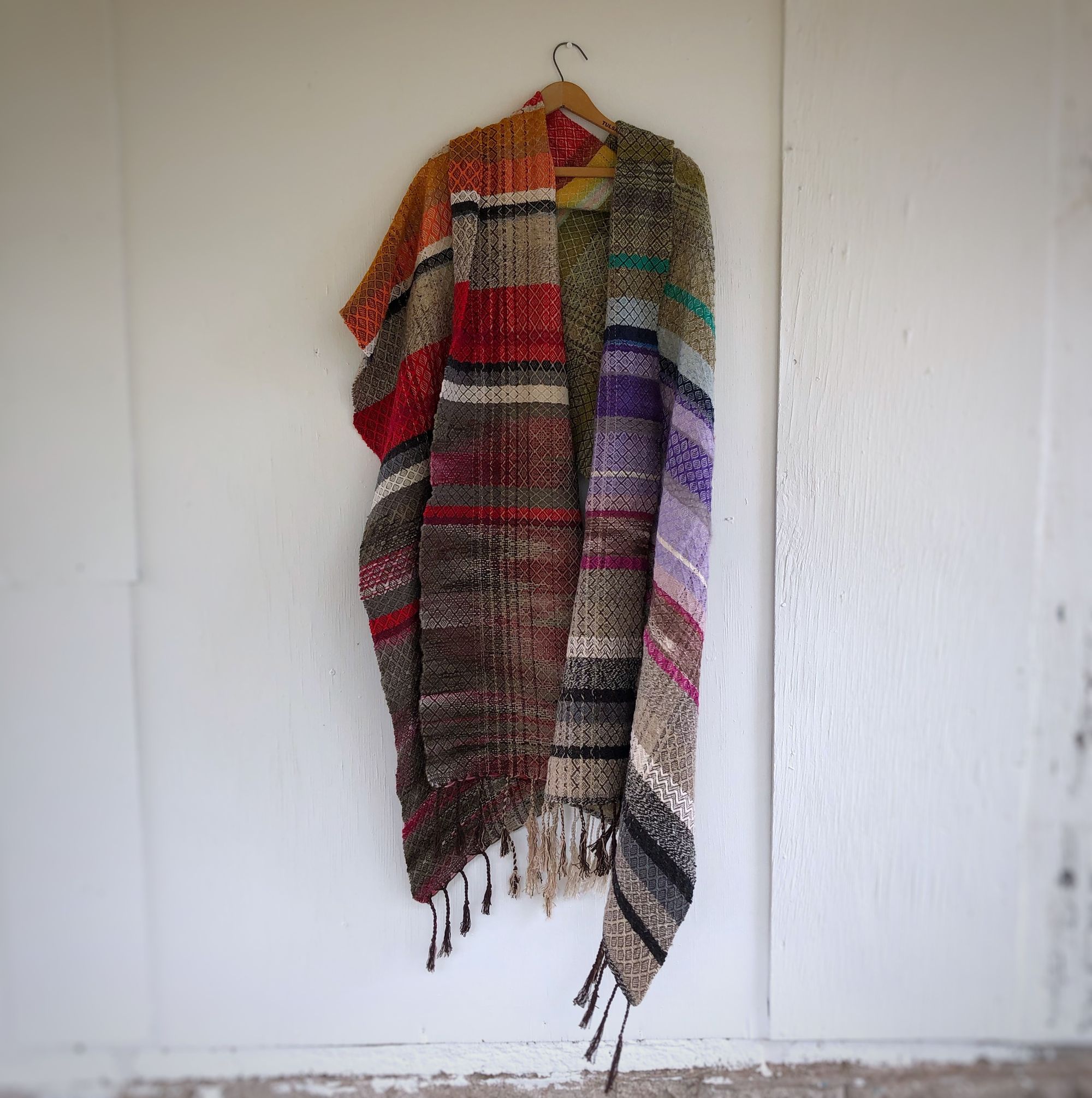 Handwoven red, white, orange, green, purple, brown and black shawl hanging on a hanger on a white wall