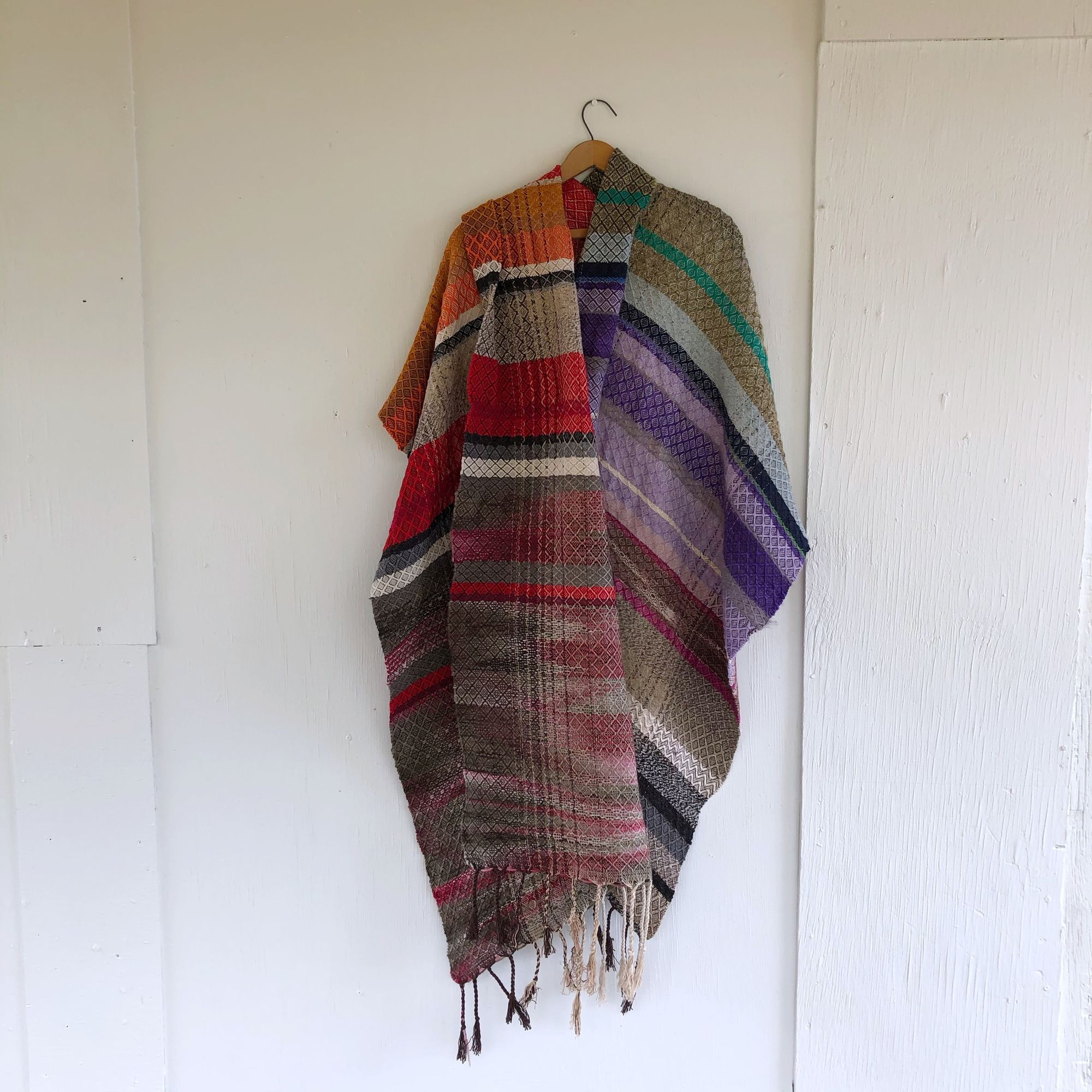 Handwoven red, white, orange, green, purple, brown and black shawl hanging on a hanger on a white wall