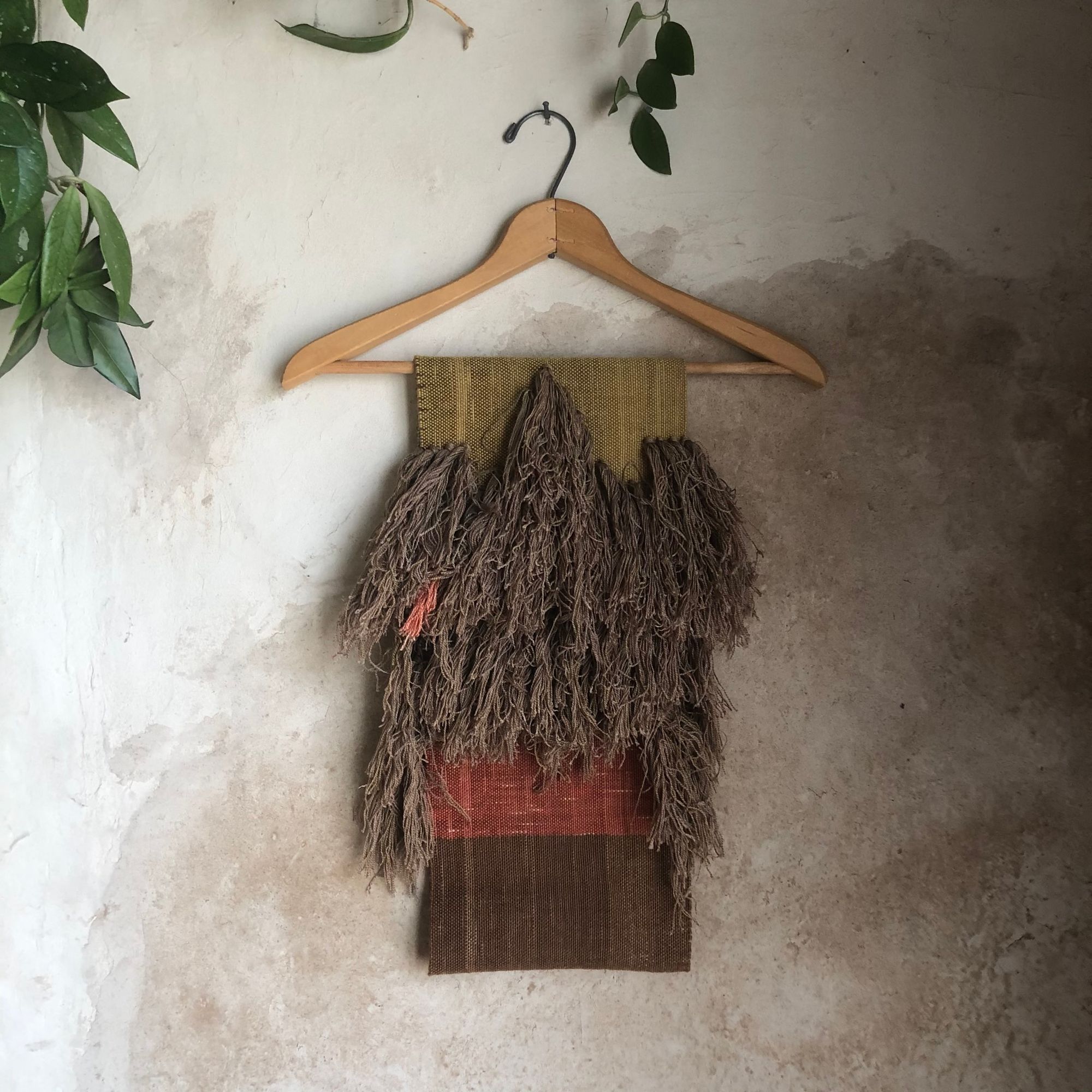 handwoven highly textured scarf that is mustard, brown, grey-blue and salmon colored, on a hanger against a wall
