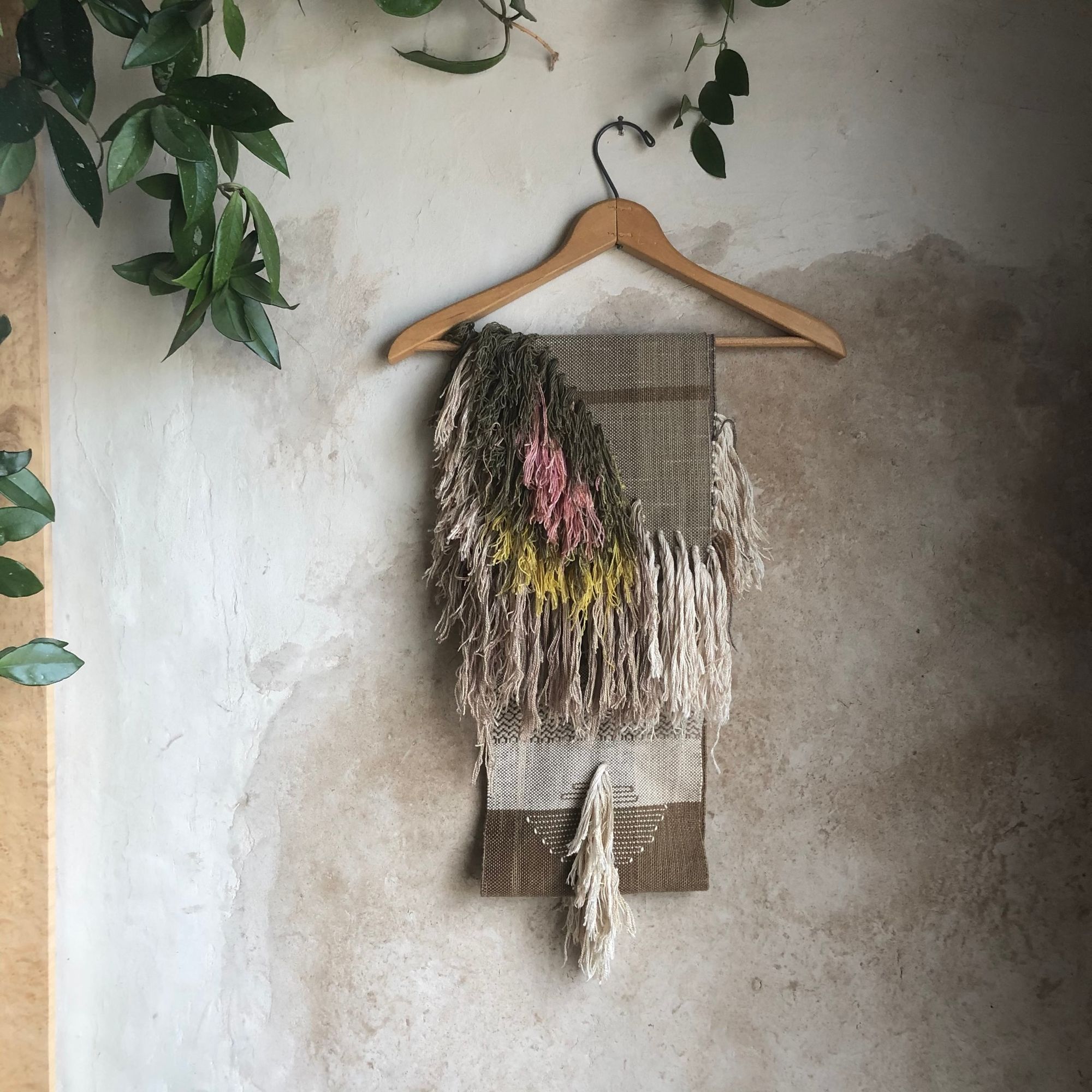 handwoven natural dye brown silk scarf with white, green, pink and yellow fringe hanging on a hanger on a white mud wall 