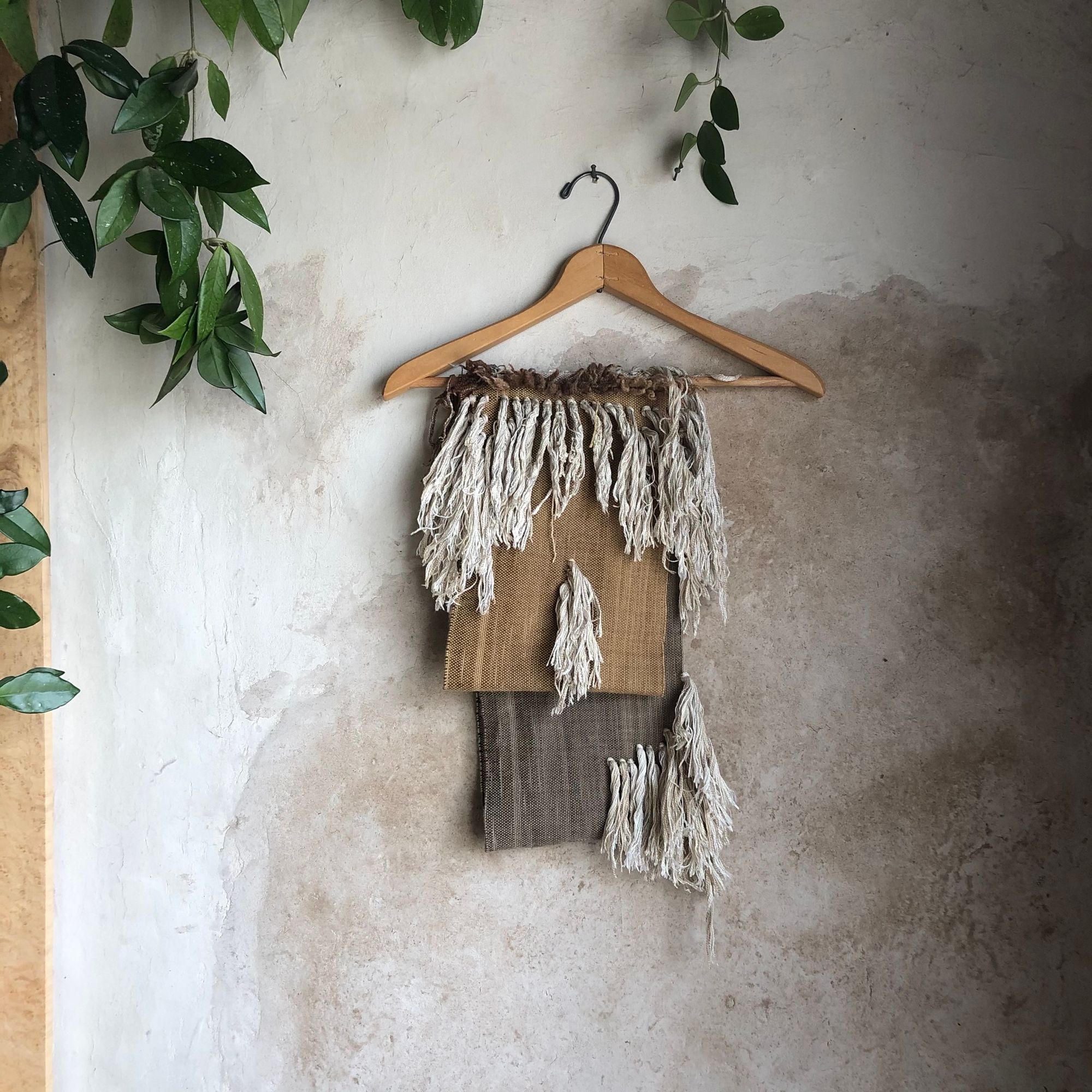 salmon colored, brown, white and grey scarf with white cashmere fringe on a hanger, hanging on a white and tan wall with a plant in the corner. 