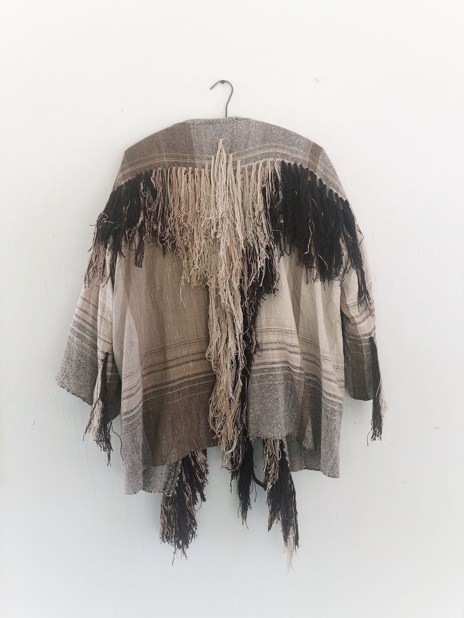 raw silk and organic cotton linen brown, tan and white Handwoven fringed Cloak hanging on a hanger on a white wall
