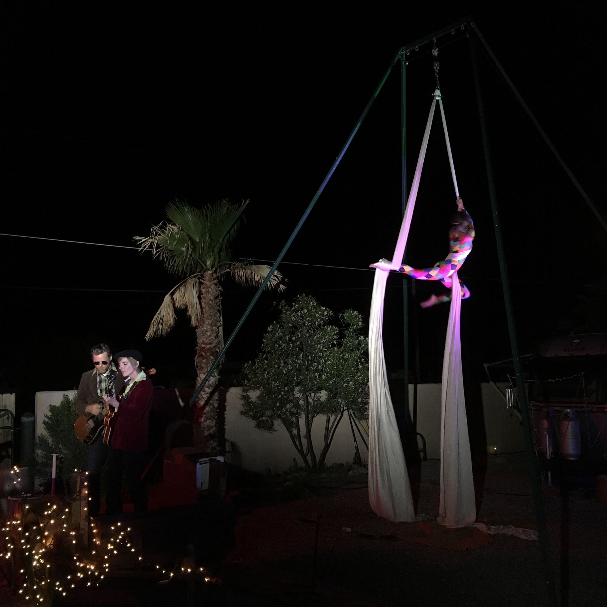 Woman wearing multicolored harlequin costume doing aerial acrobatics at night while two people play music in front.