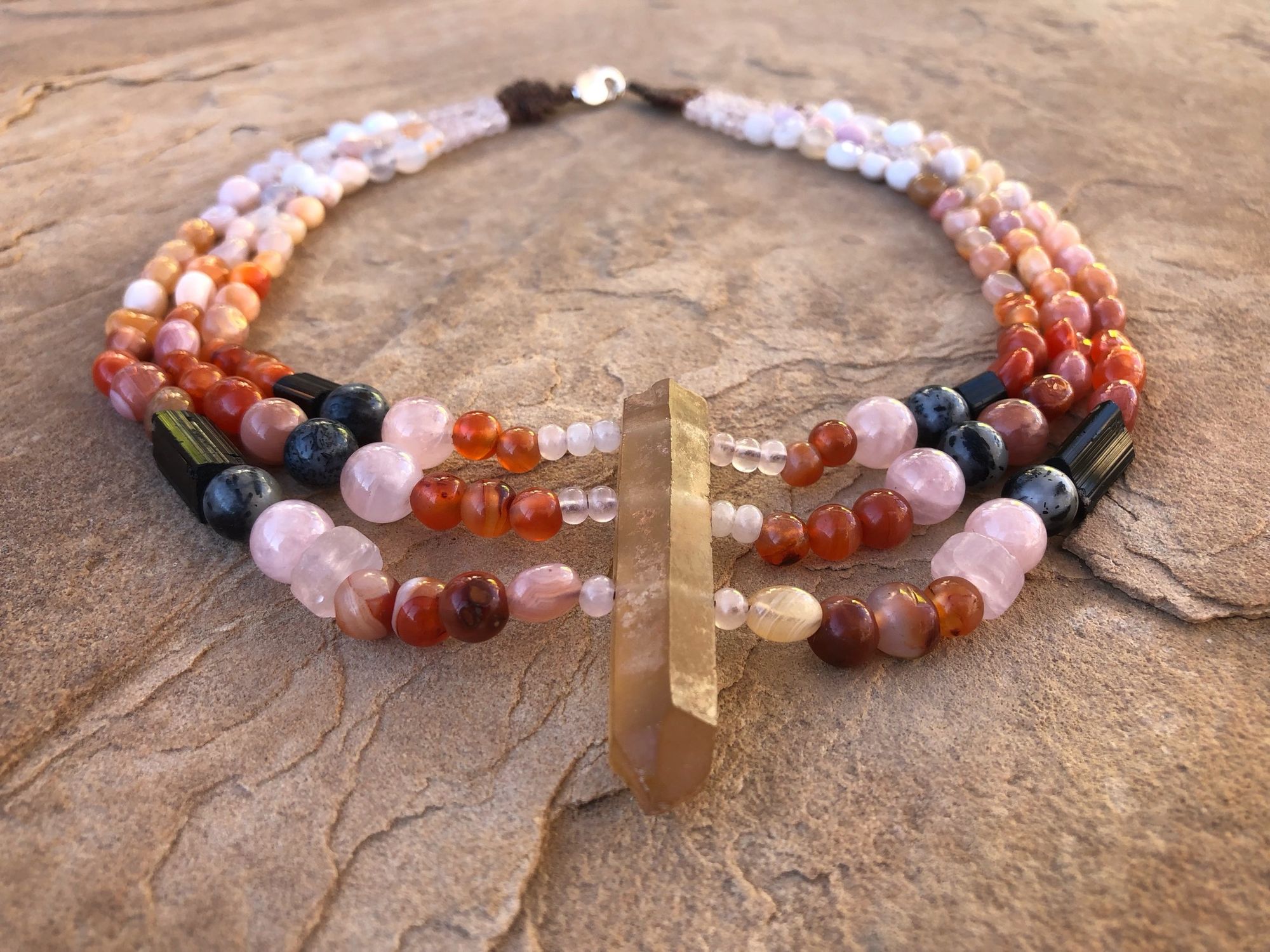 Necklace laying on tan sandstone that has citrine yellow-brown, rose quartz pink, black tourmaline and red agate beads