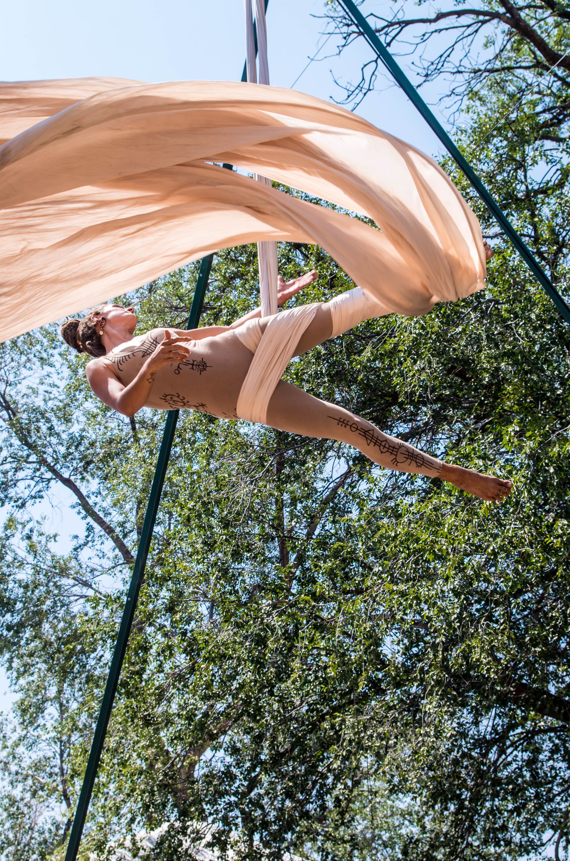 Woman in tan leotard going aerial acrobatics on white fabric next to a stage and large tree at a music festival. 