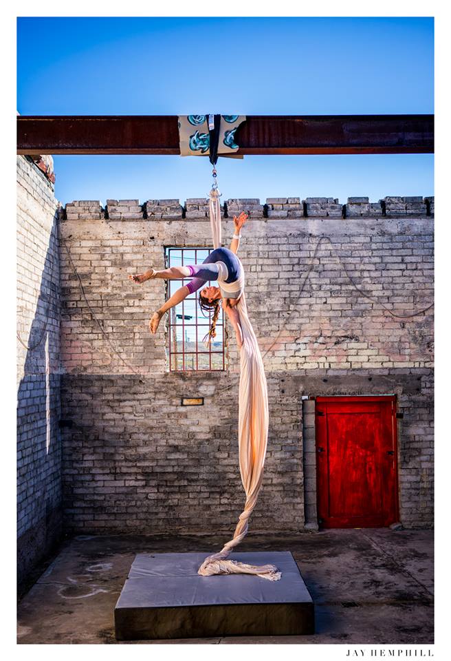 Woman in a grey shirt dong aerial acrobatics in front of a grey brick wall with a red door. 