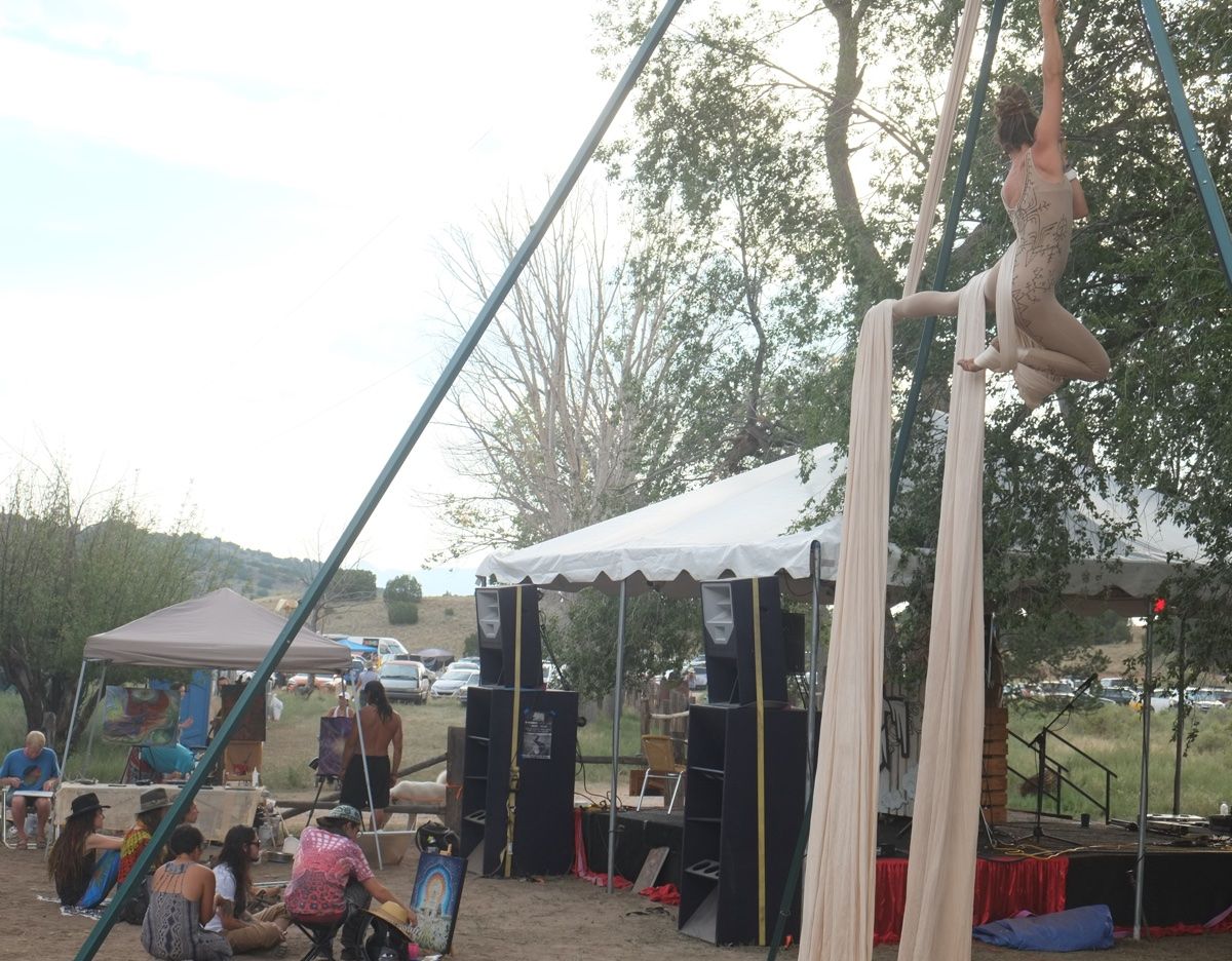 Woman in tan leotard going aerial acrobatics on white fabric next to a stage and large tree at a music festival. 