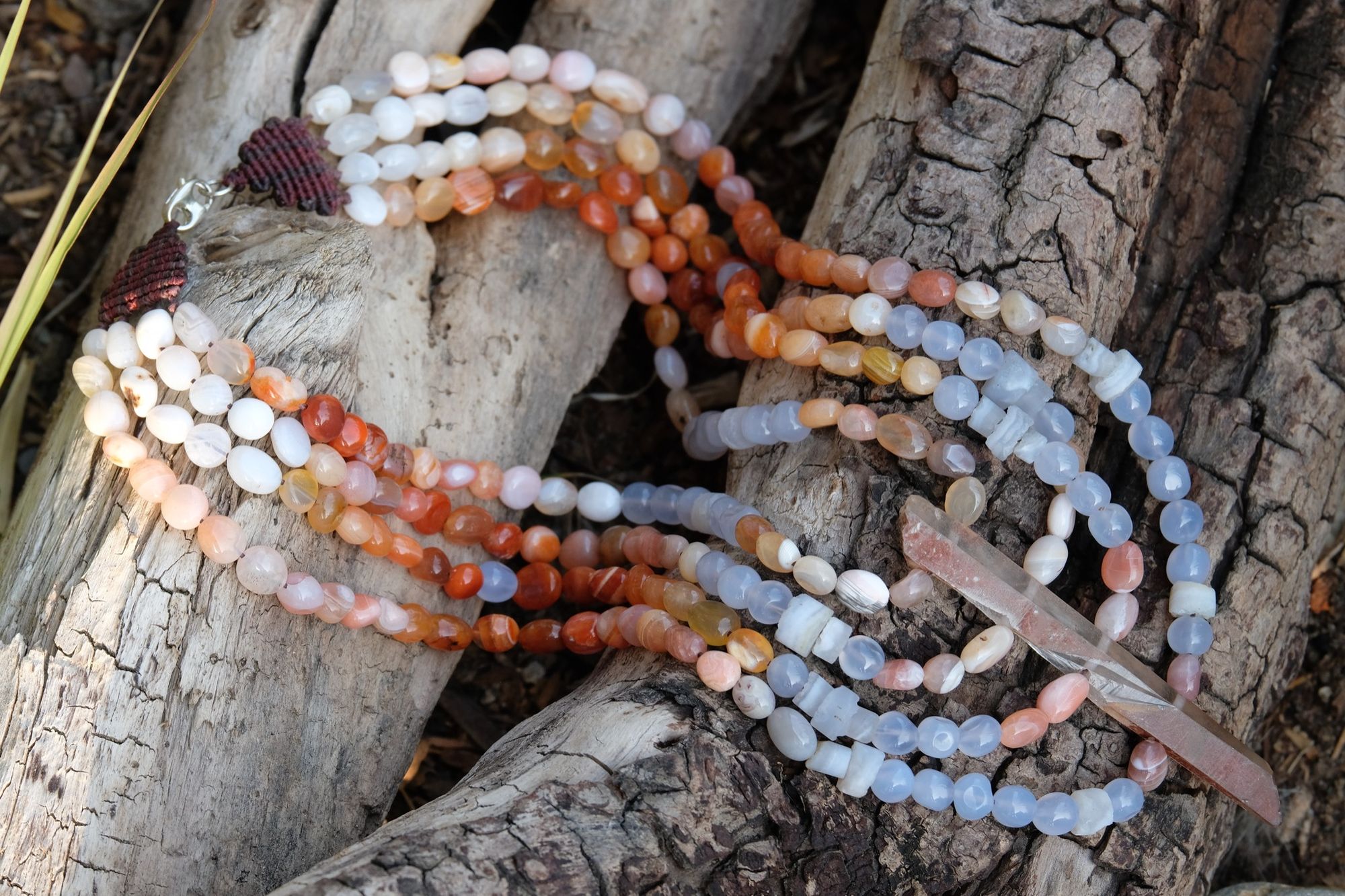 Detail of clear quartz point, red-pink-white graded Botswana agate and blue chalcedony necklace laying on a log